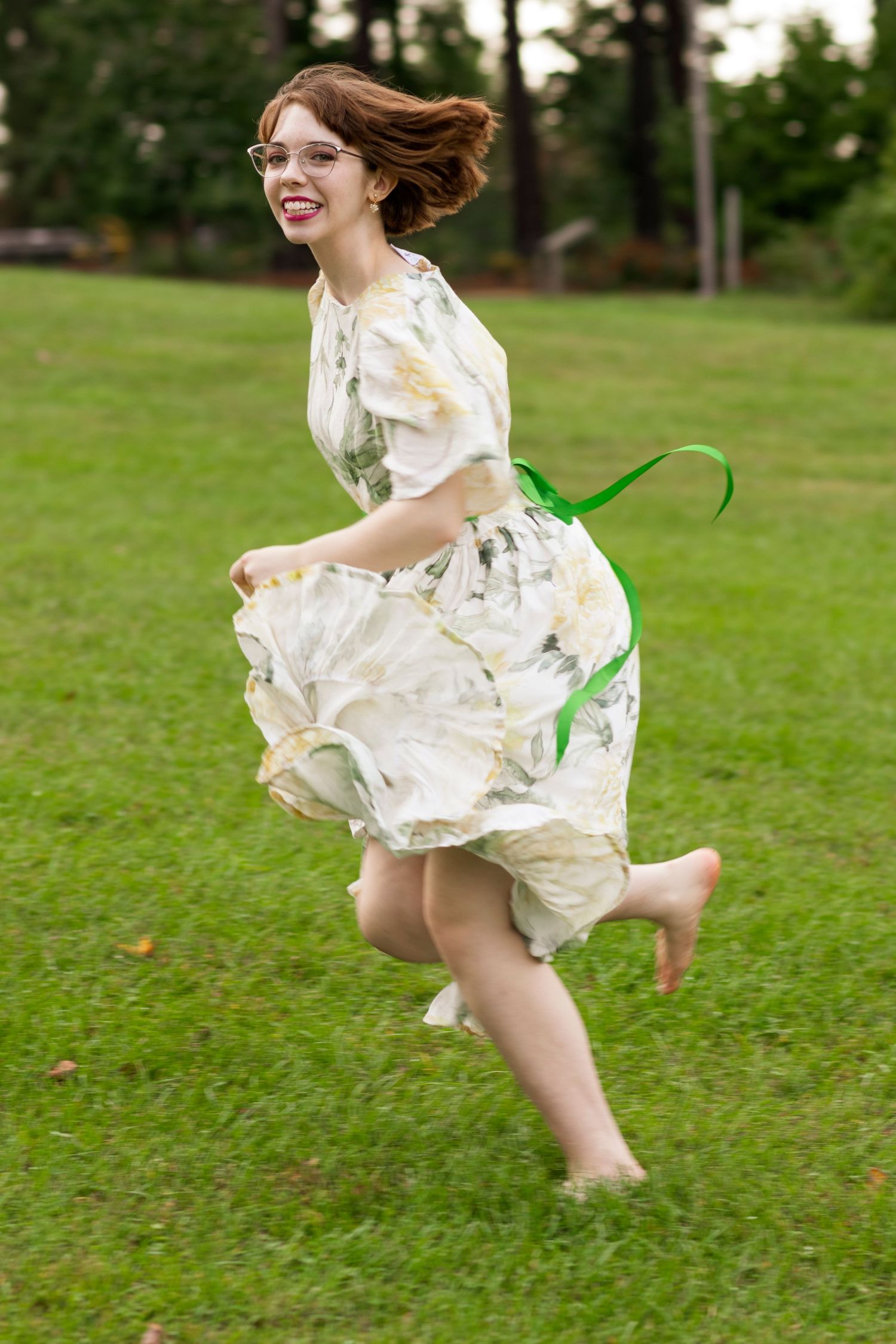A joyful figure in a flowy white dress leaps barefoot across a green lawn on a summer day.