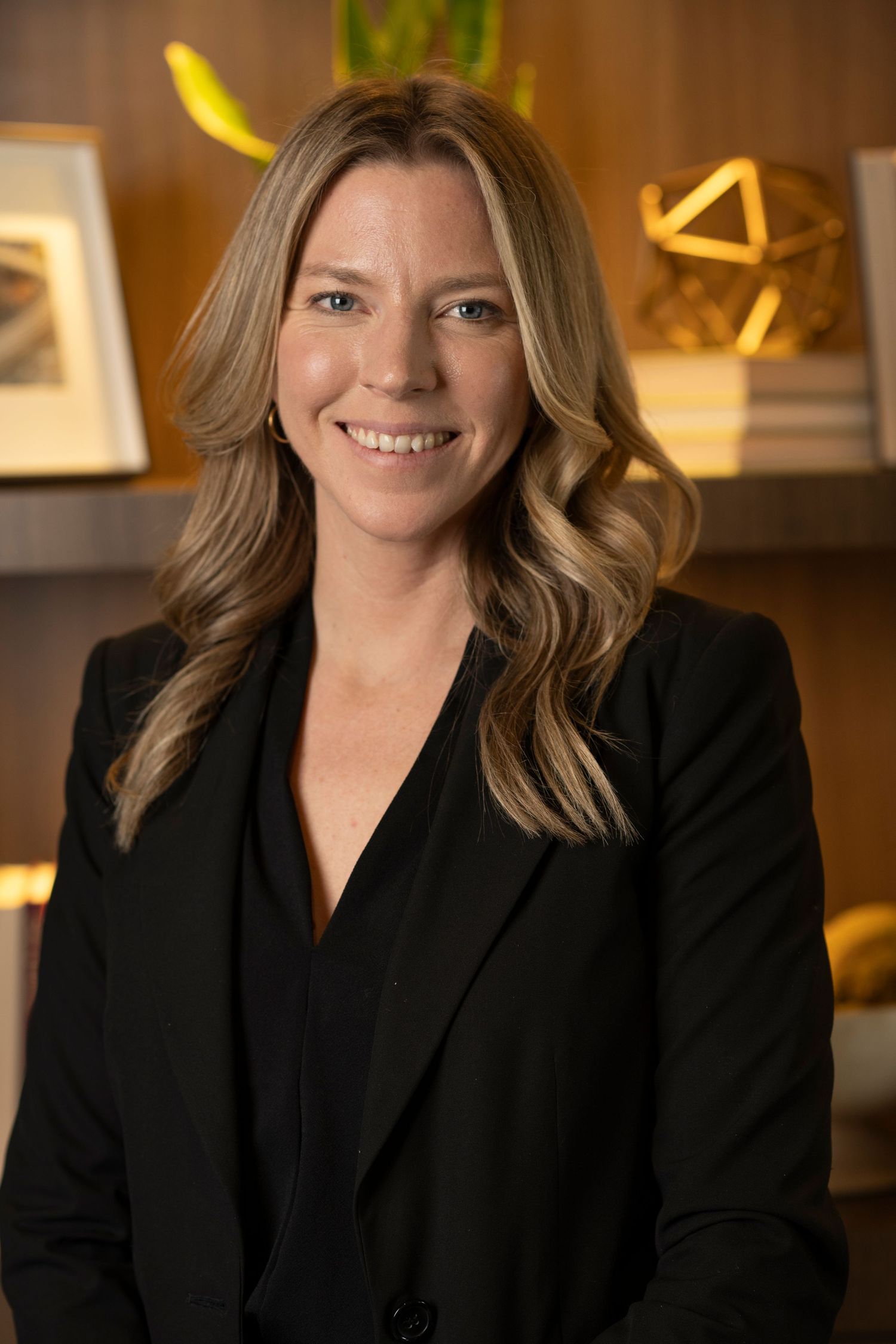 Corporate headshot of person in black blazer with wavy hair against warm office background.
