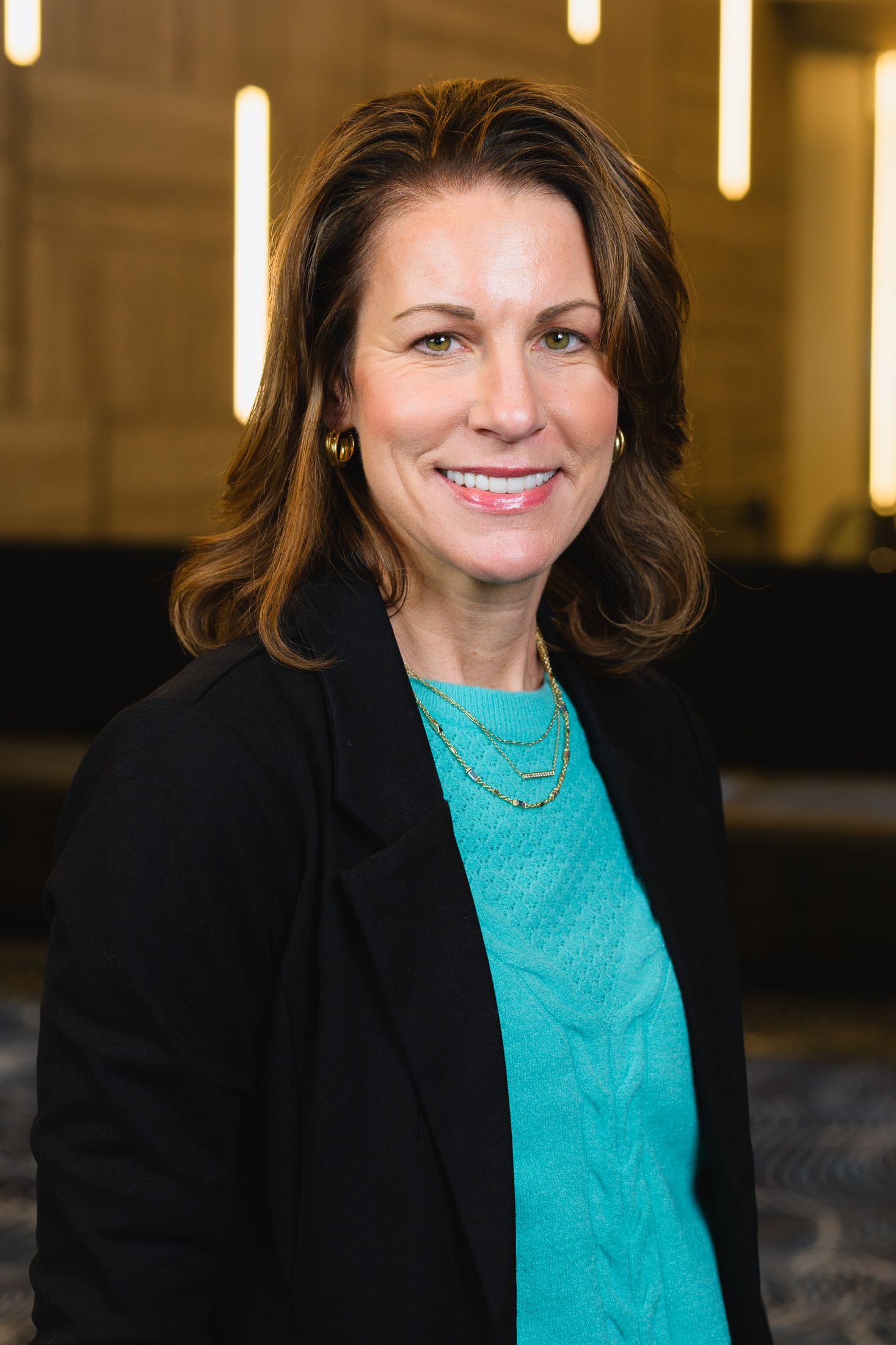 Professional portrait of someone in a black blazer and turquoise sweater with warm background lighting.