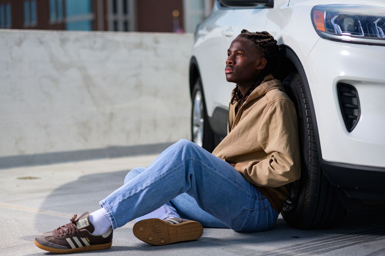 Young Black man in casual outfit sitting on ground leaning against white SUV tire outdoors on sunny day.