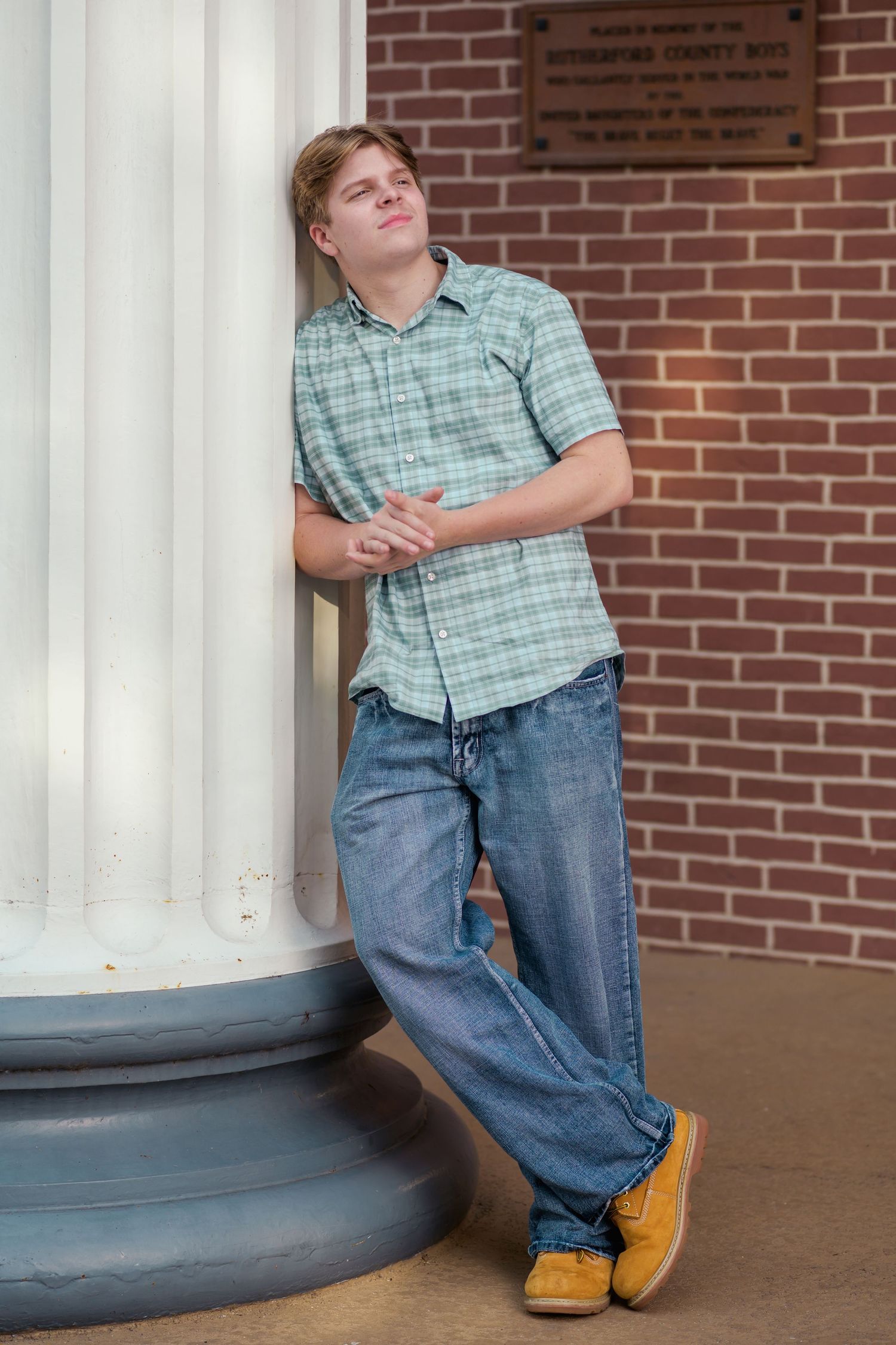 Young man in plaid shirt and jeans leaning against white column on brick building exterior.