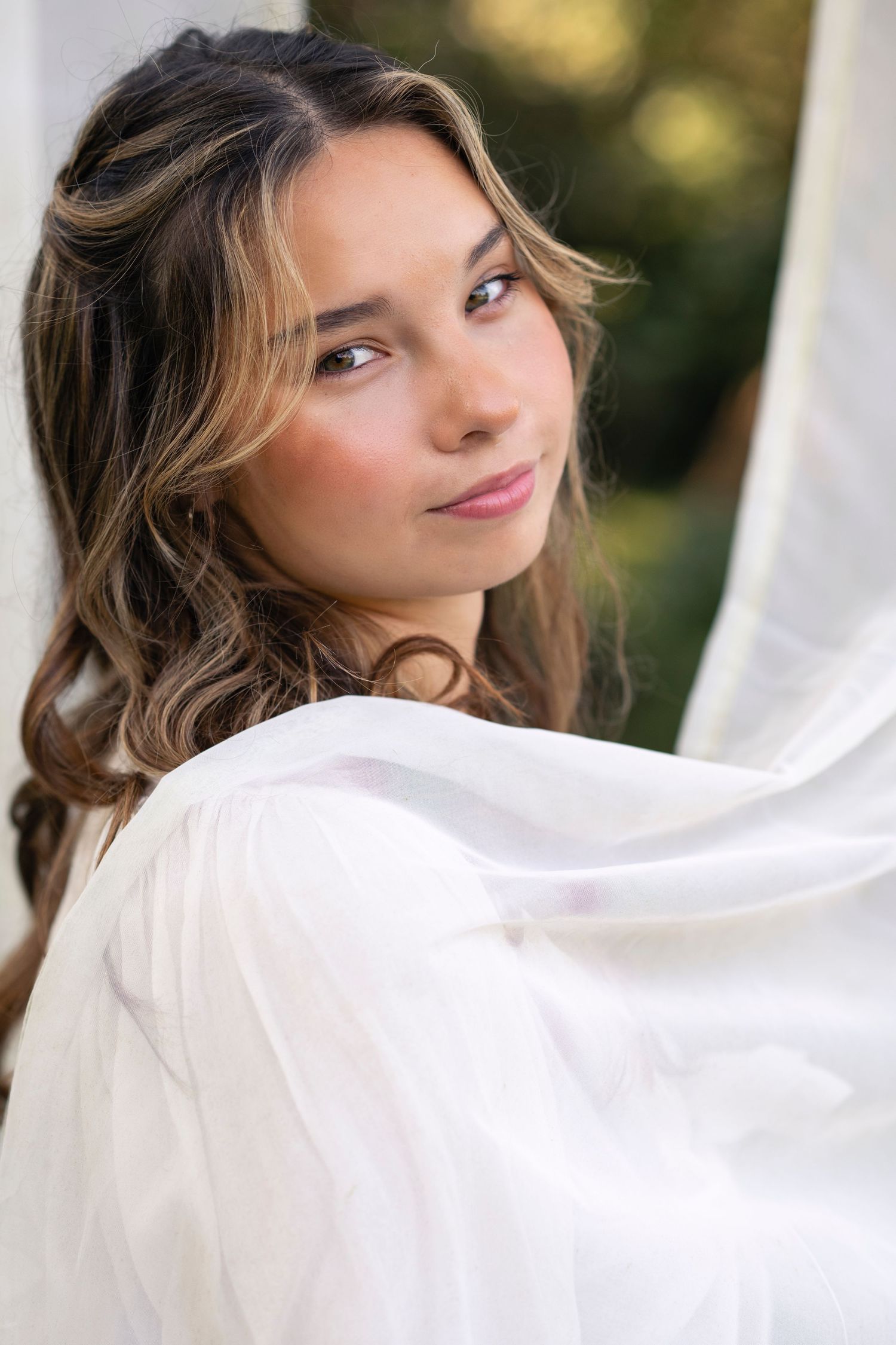 A portrait photo of someone with wavy brown hair and highlights wearing a white top in soft natural lighting outdoors.