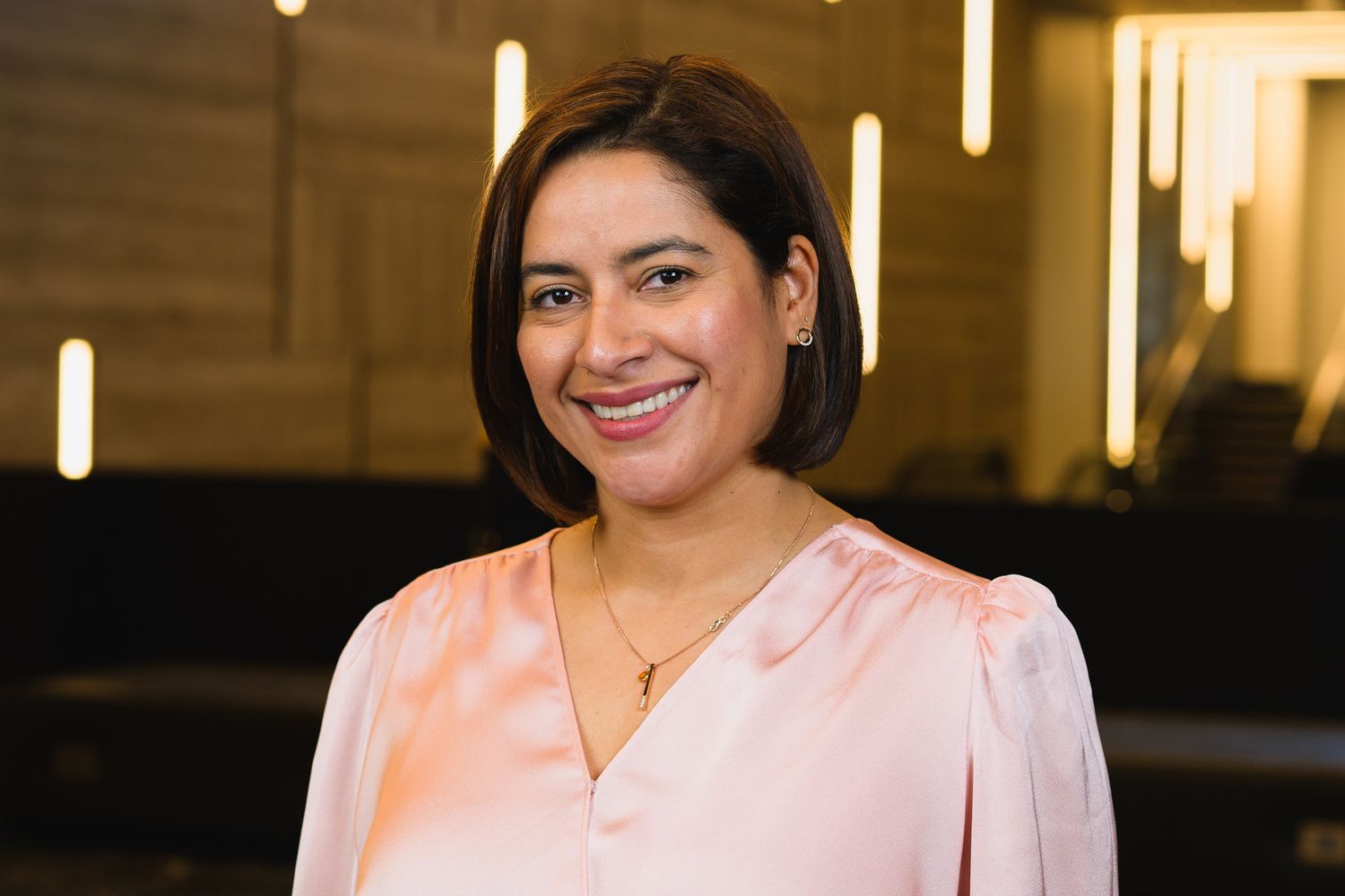 Professional portrait of someone in a pink blouse smiling against warm interior lighting.