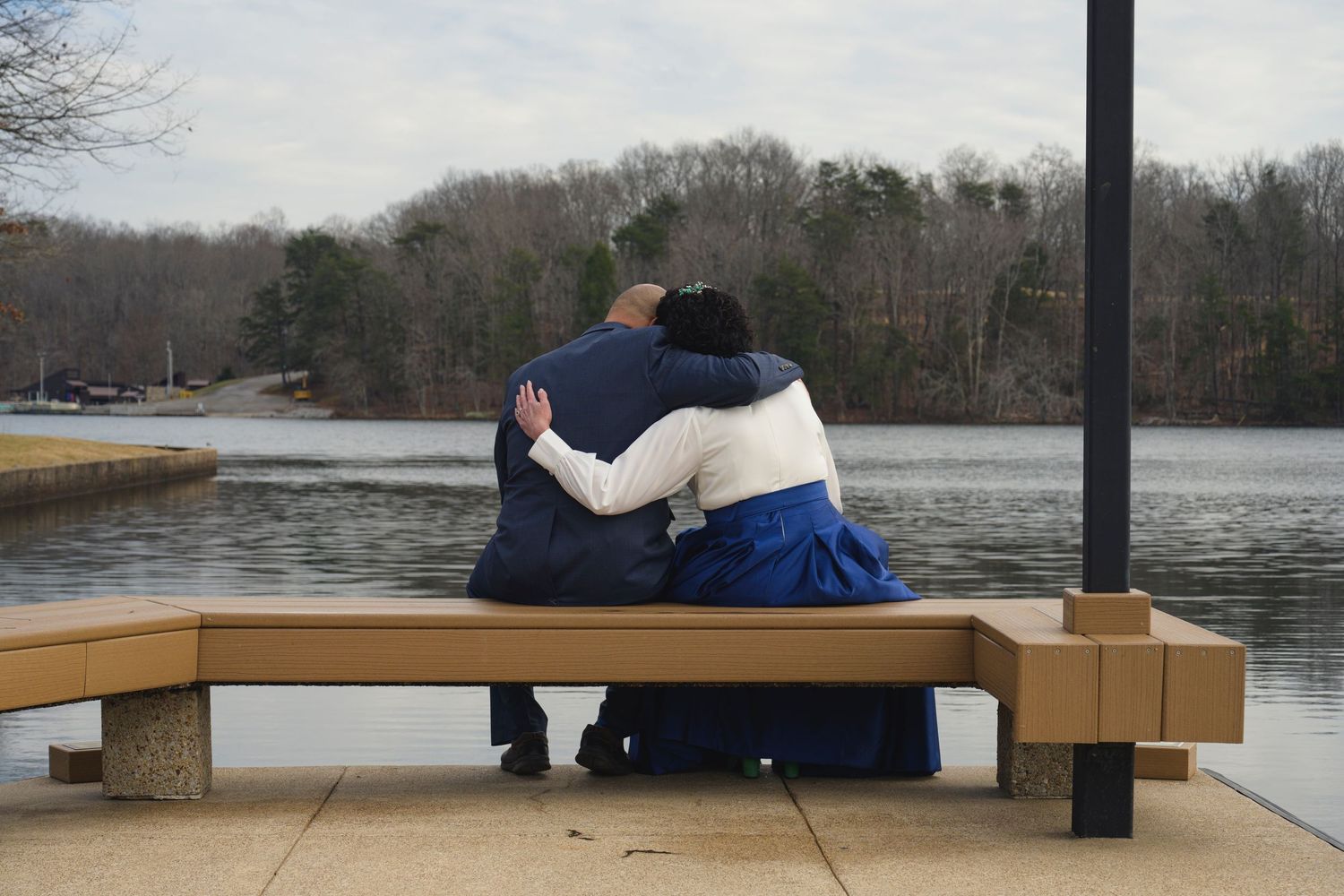 Two people sit on a wooden bench overlooking a serene lake surrounded by bare winter trees.