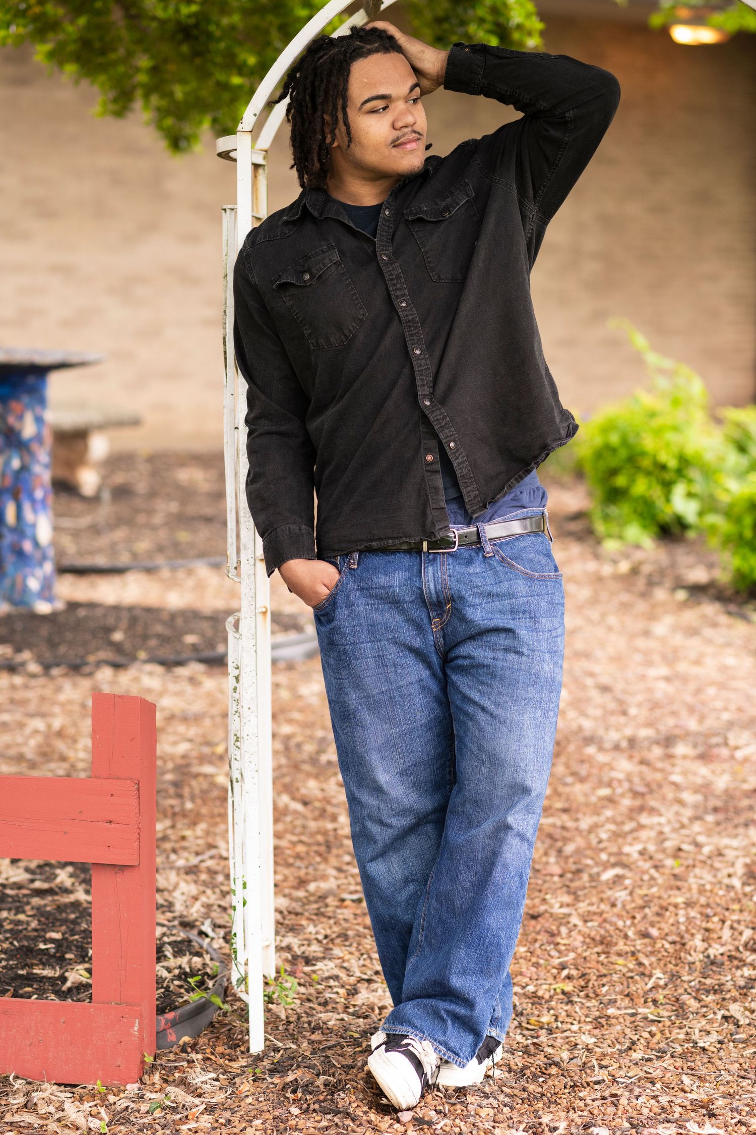 Person wearing black jacket and blue jeans poses casually against white post in outdoor garden setting.