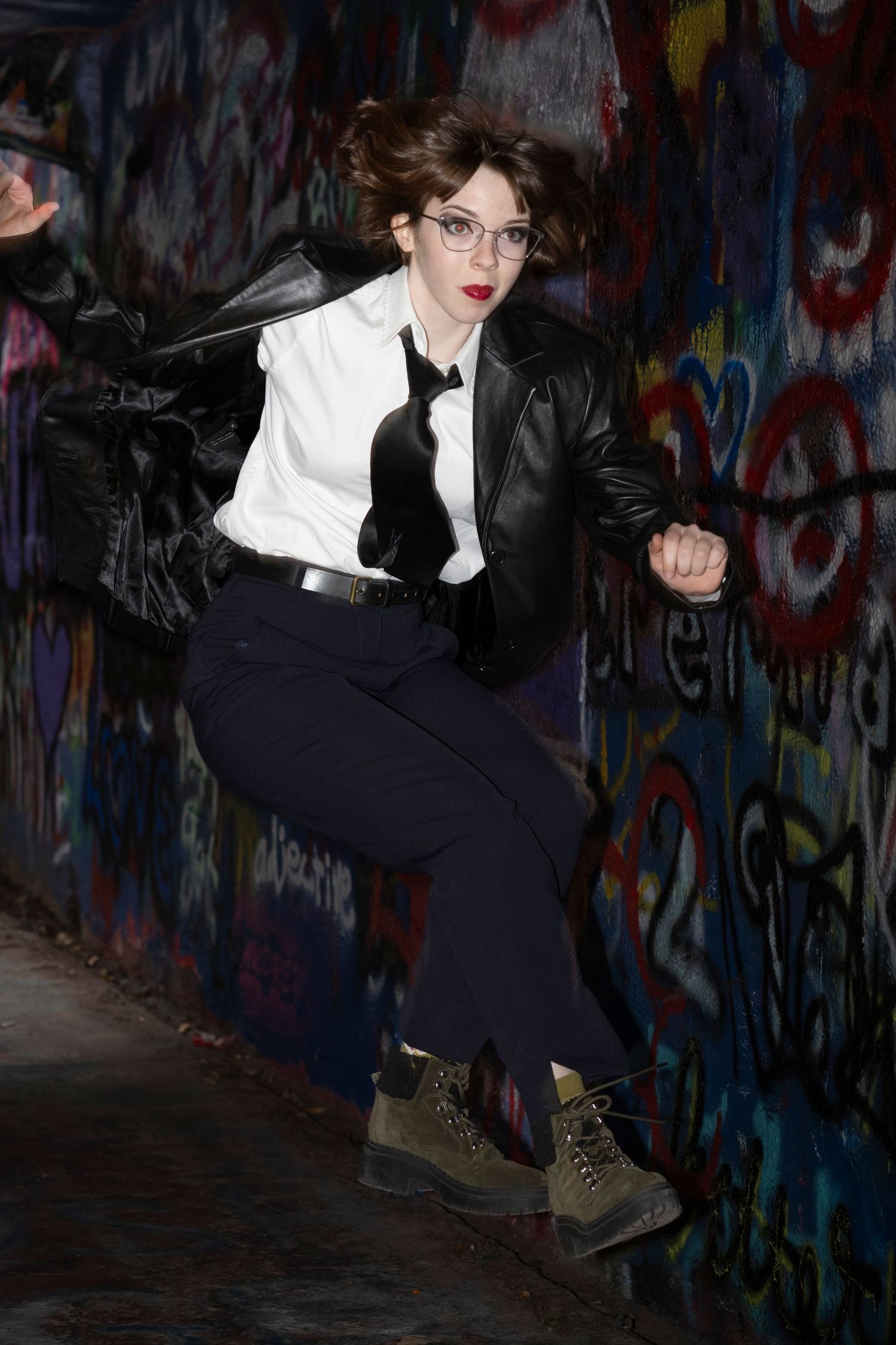A person in black and white attire performs a dynamic kicking pose against a graffiti-covered wall.