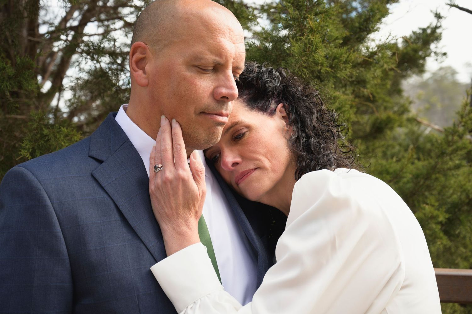 A tender romantic moment between a couple in formal attire standing outdoors near evergreen trees.