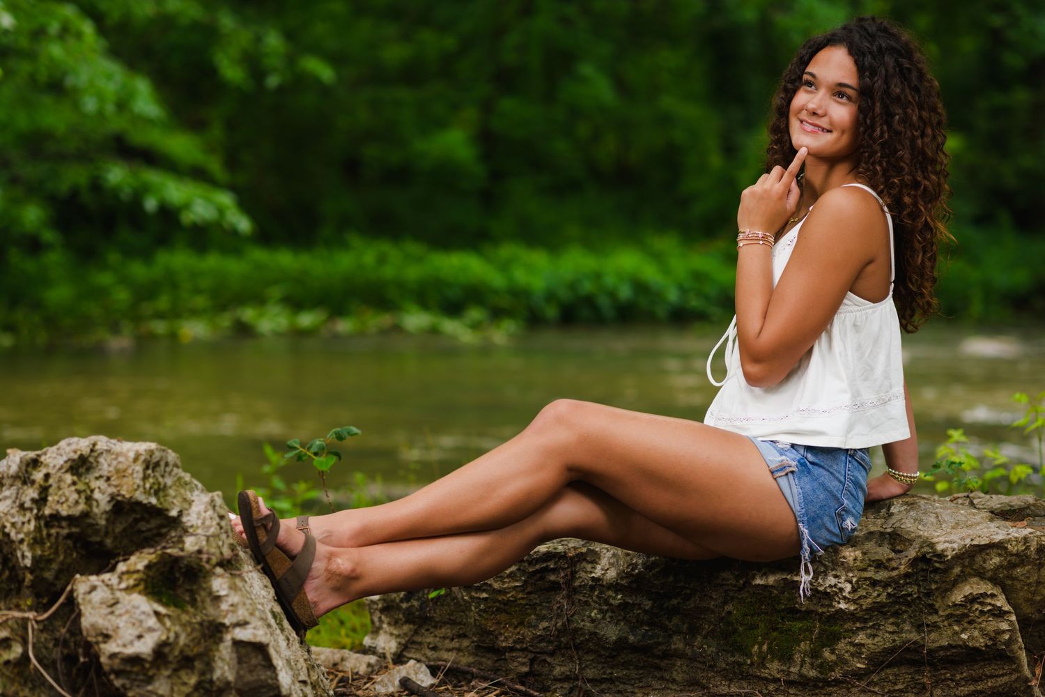 Young woman with curly hair in white top and denim shorts sitting on rocks by a river in a lush green forest.