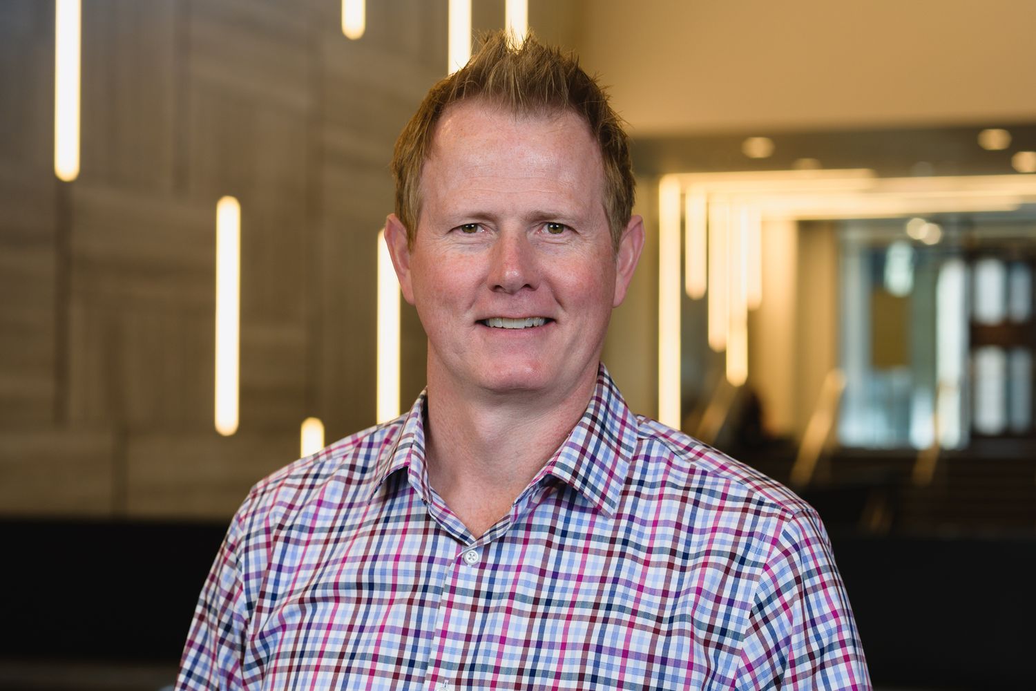 Professional headshot of someone wearing a blue and white checkered shirt and navy blazer in an office setting.