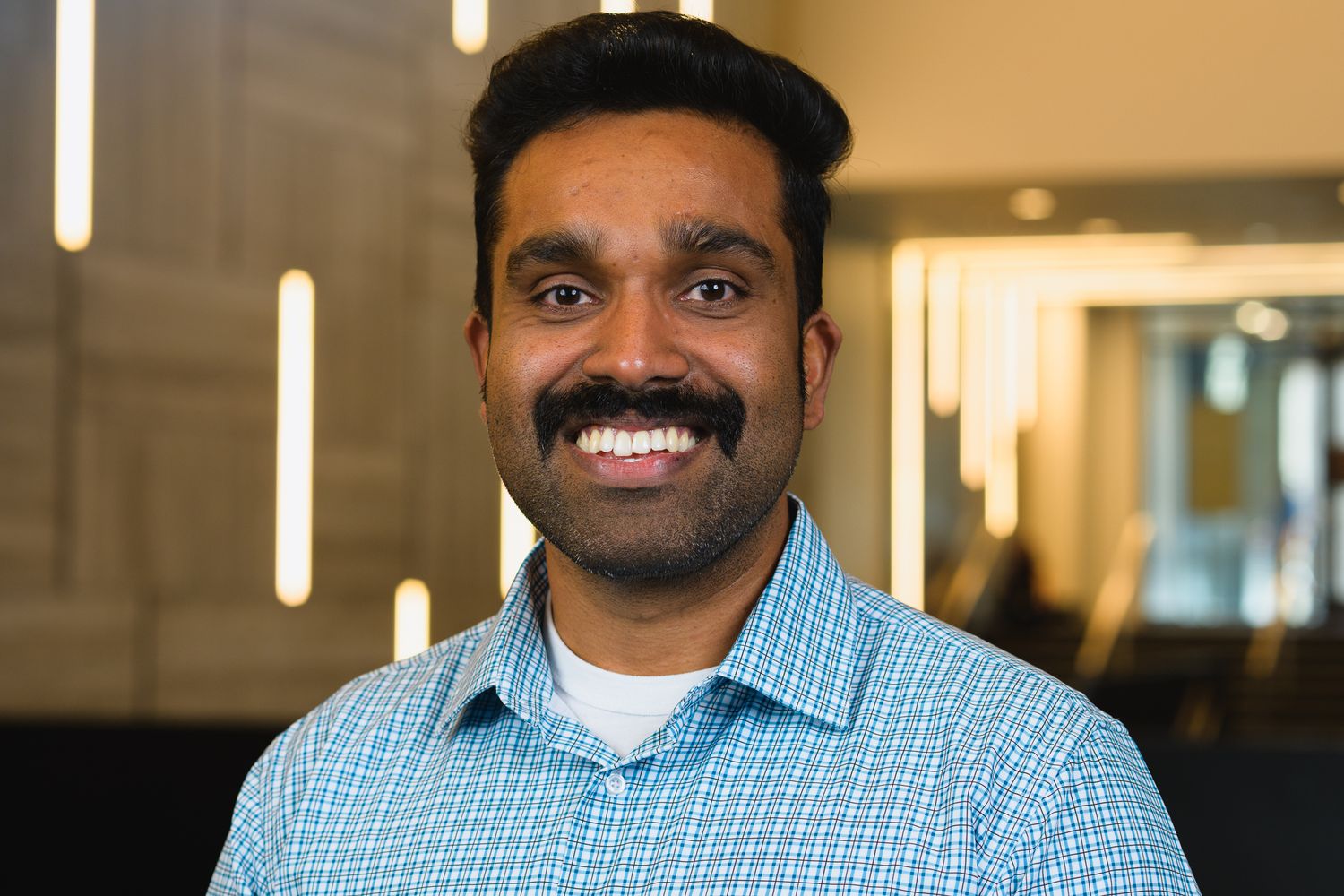 Professional headshot of person in light blue shirt smiling against office background.