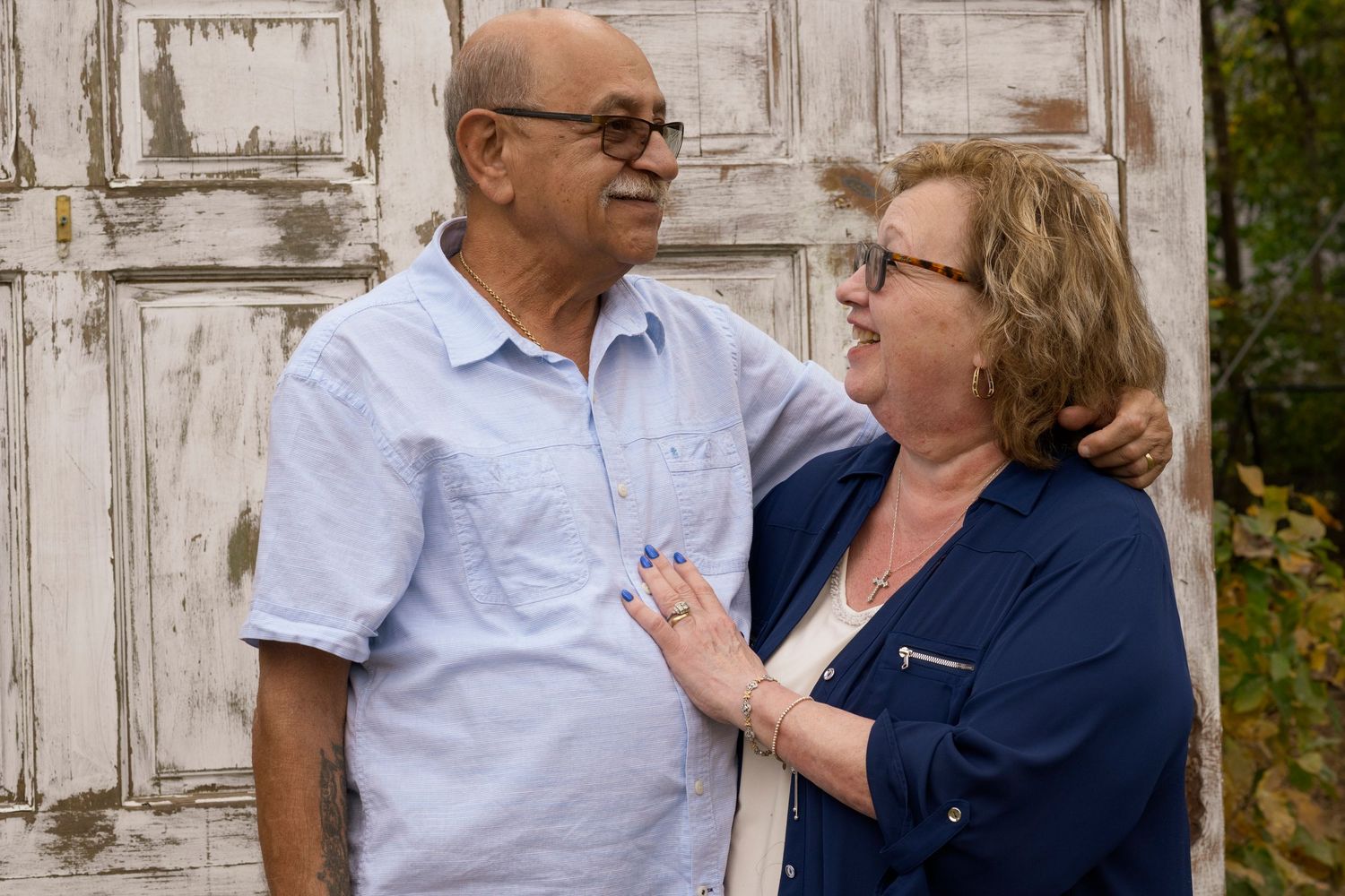 A couple embraces lovingly in front of a weathered white door during an outdoor photoshoot.