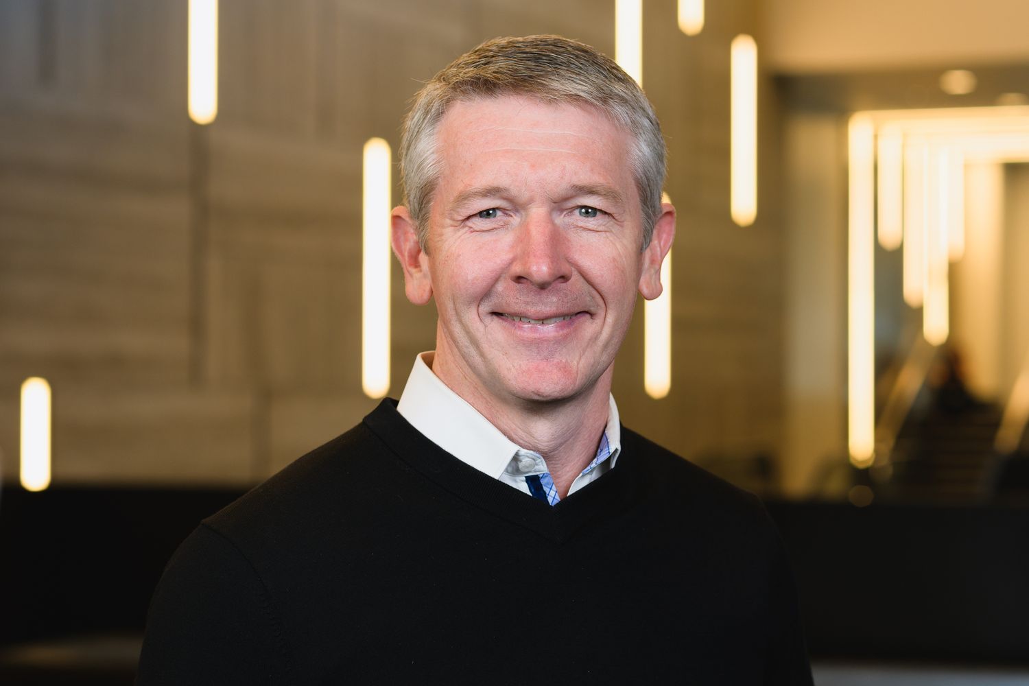 Professional portrait of person in dark jacket against modern office backdrop.