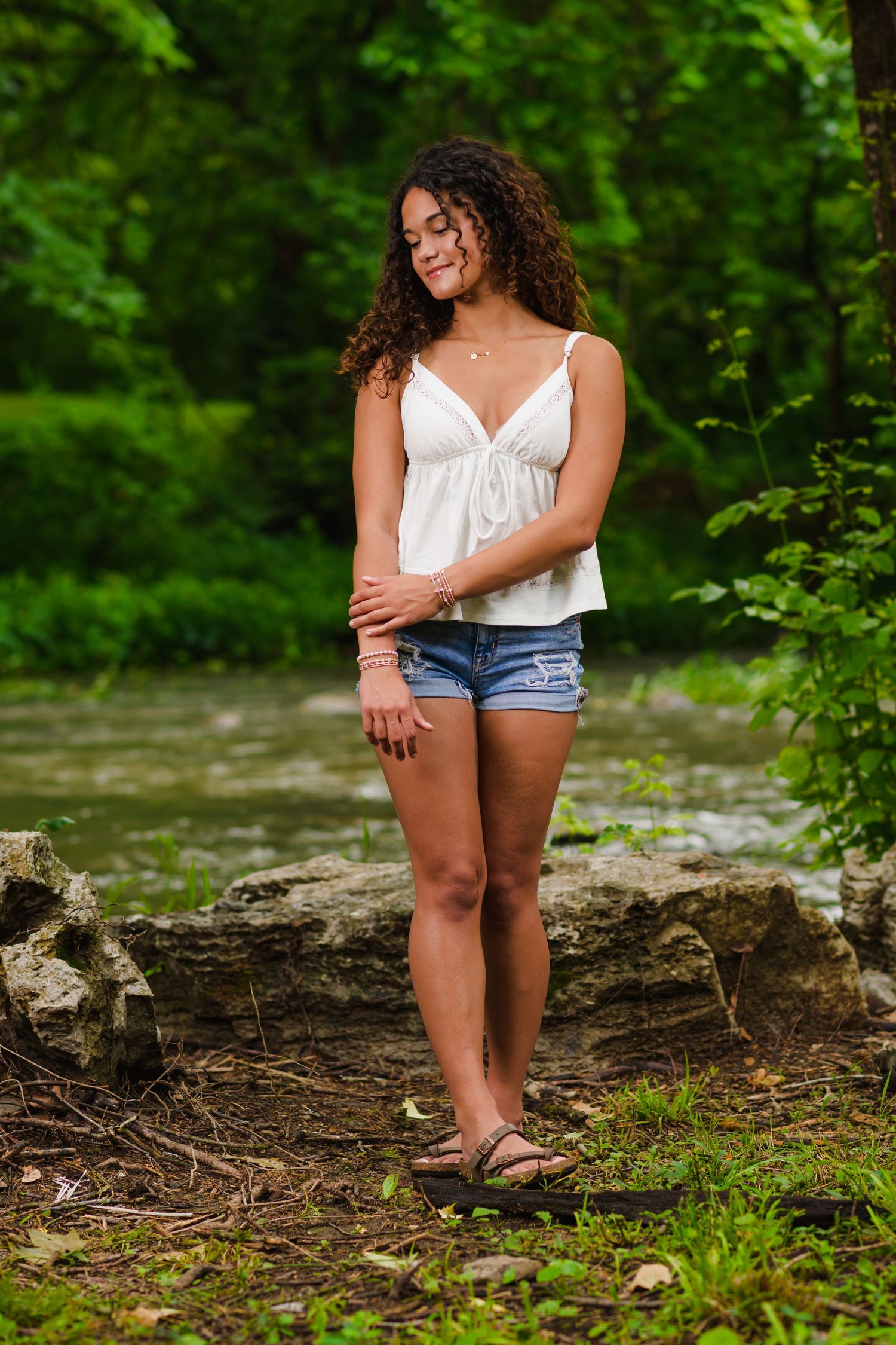 Young woman with curly hair in white top and denim shorts standing near rocks in a lush green forest.
