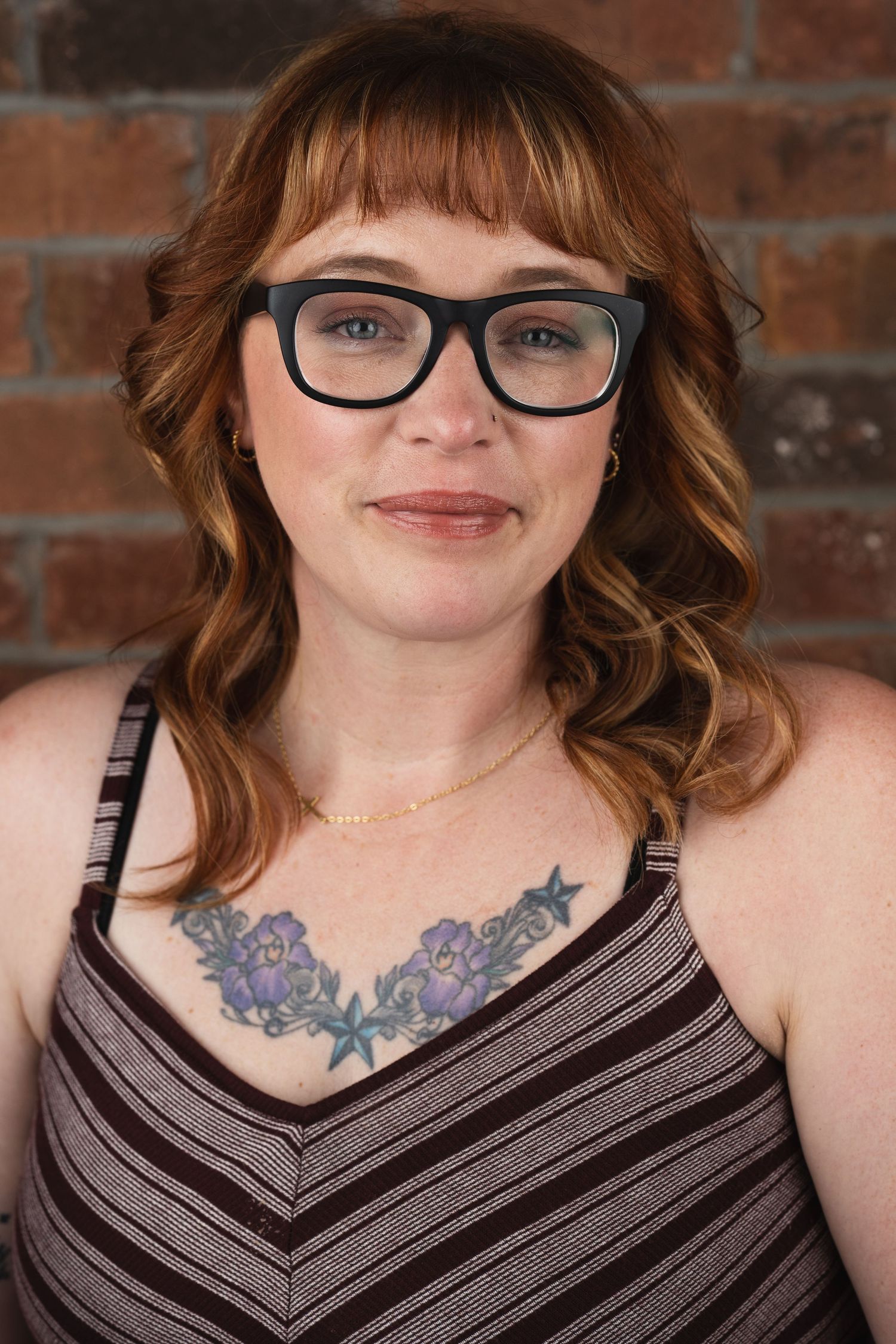 Portrait against brick wall showing chest tattoo and striped top in warm lighting.