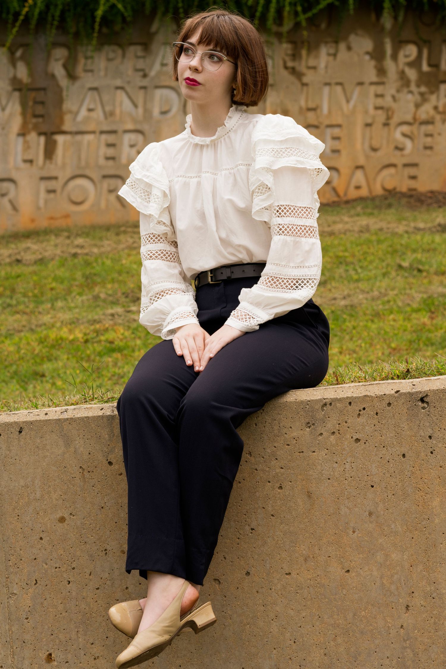 Someone wearing a white lace blouse and black pants sits casually on a concrete ledge outdoors.