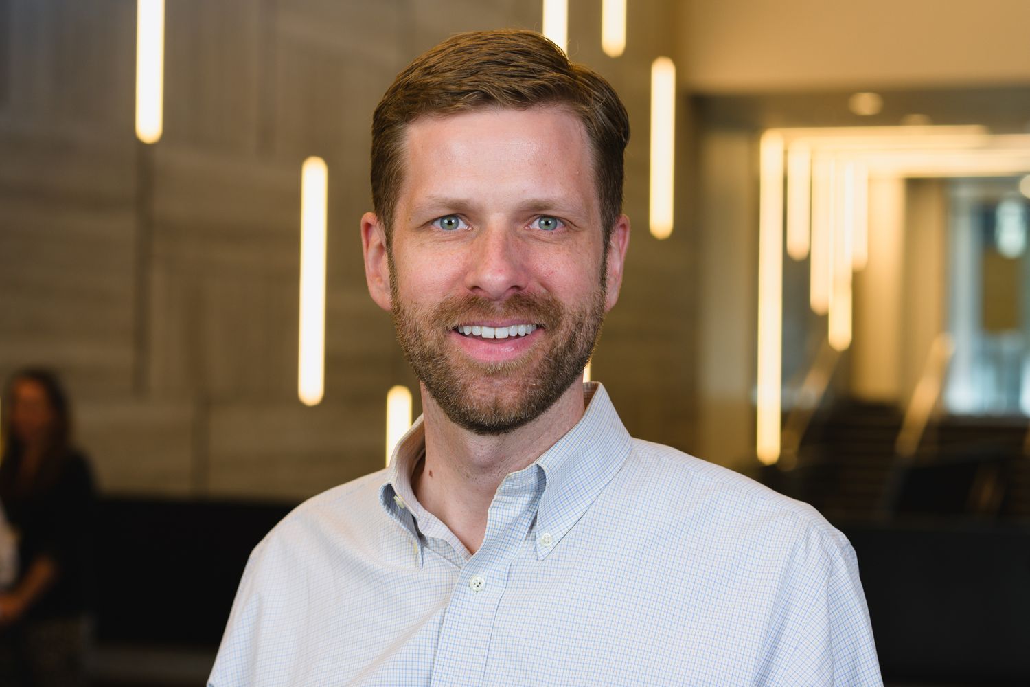 Professional headshot of person in white button-down shirt against modern office backdrop.
