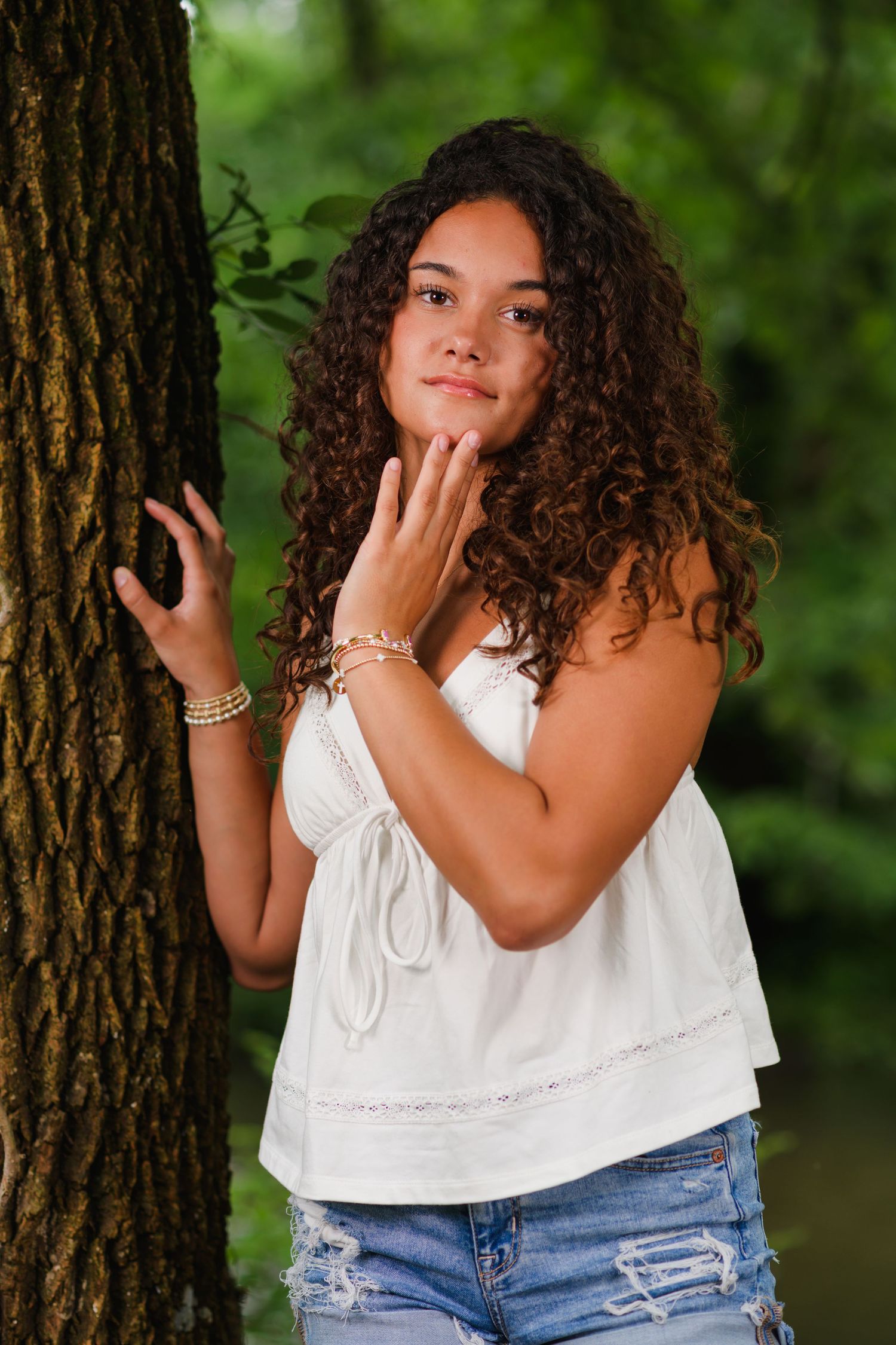 Young woman with curly hair in white top and jeans poses beside a tree in a lush green outdoor setting.