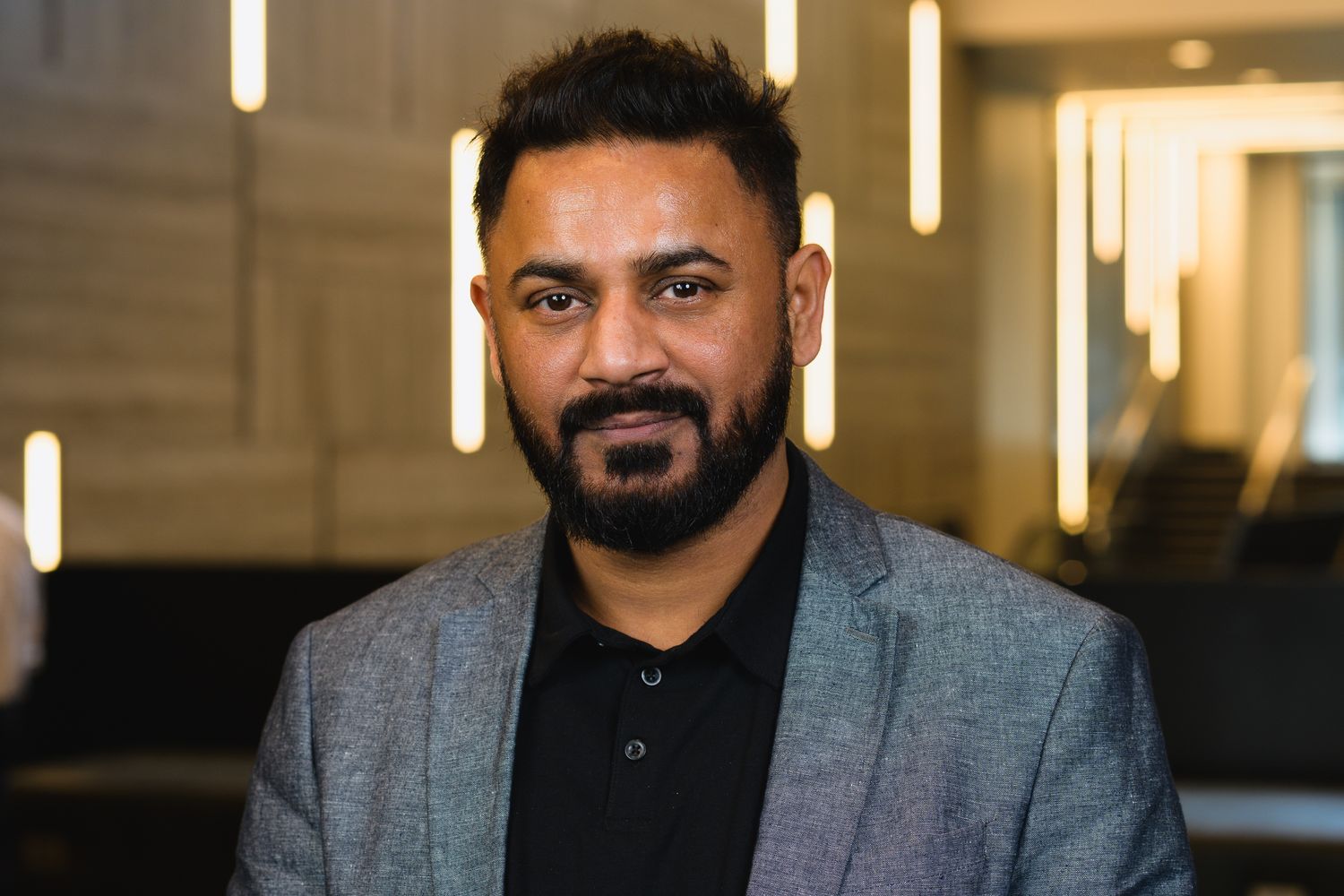 Professional portrait of person in grey blazer and black shirt against modern office setting.