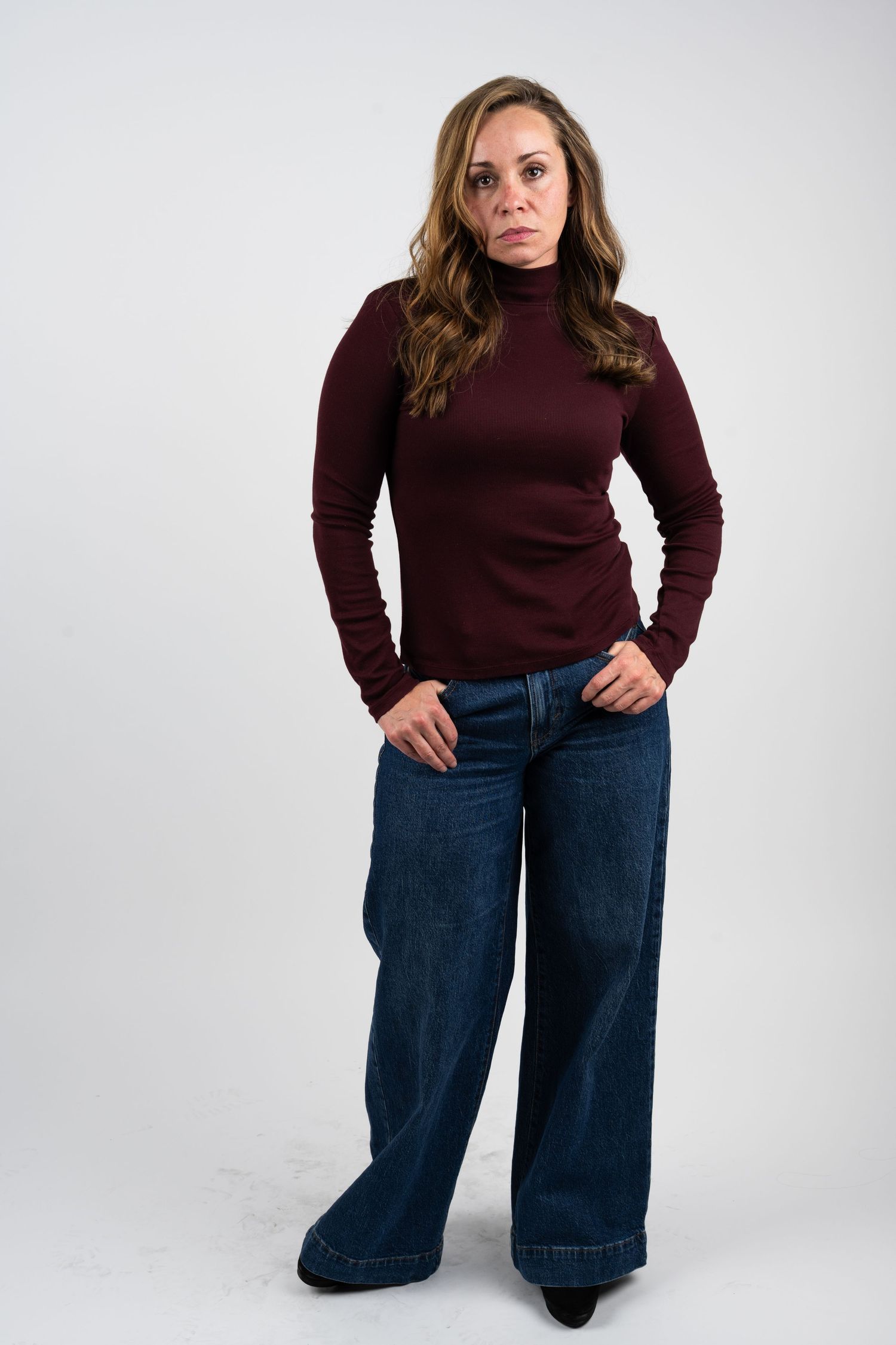 Model wearing burgundy sweater and wide-leg jeans poses in studio.