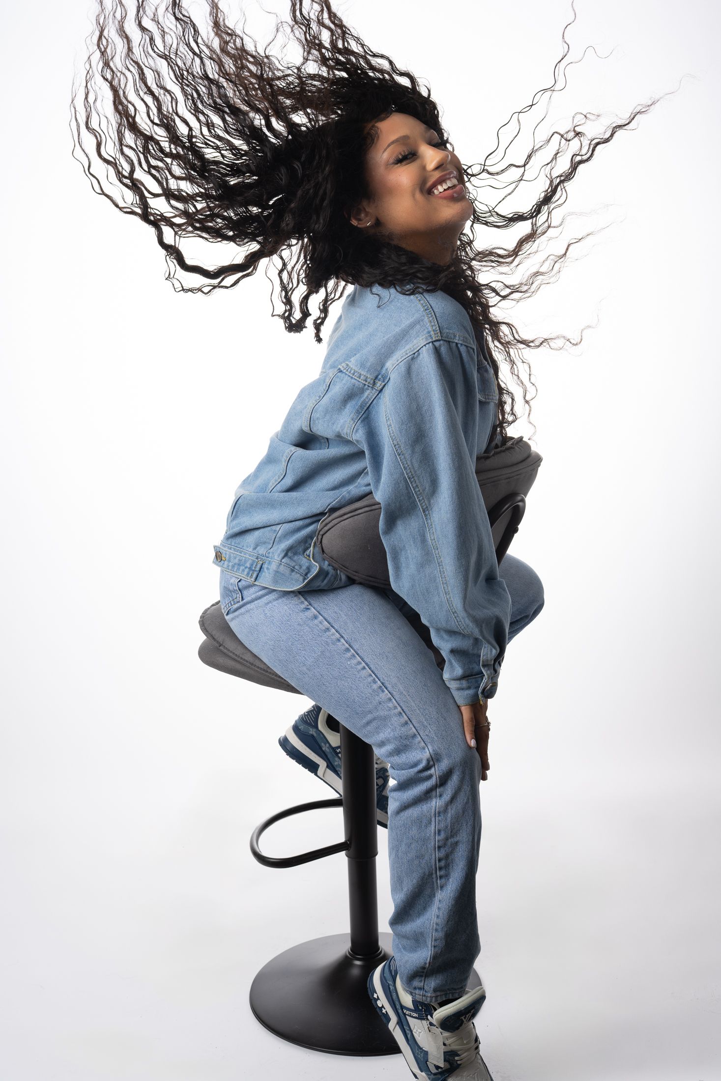 Person with long wavy hair sitting on stool in denim outfit with dramatic hair motion.