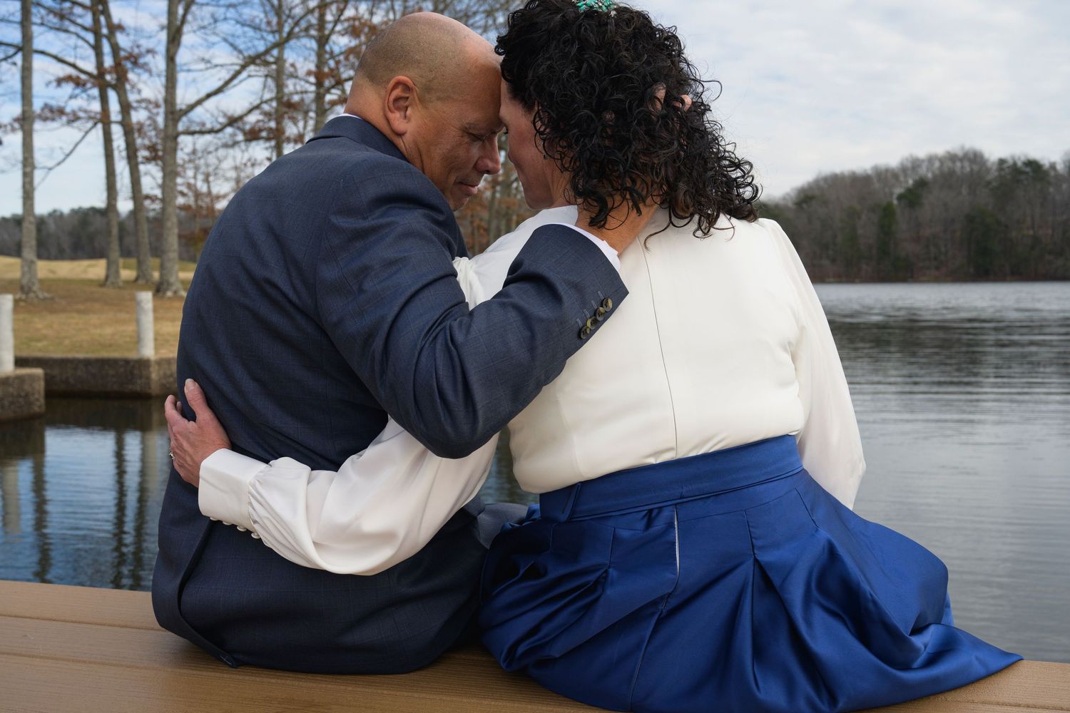 A couple sits close together on a wooden deck overlooking a serene lake surrounded by pine trees.