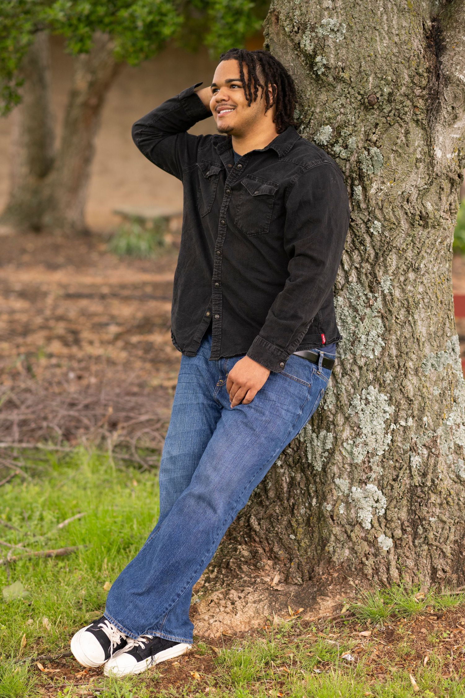 Person in black shirt and blue jeans sits casually against a large tree trunk in an outdoor setting.