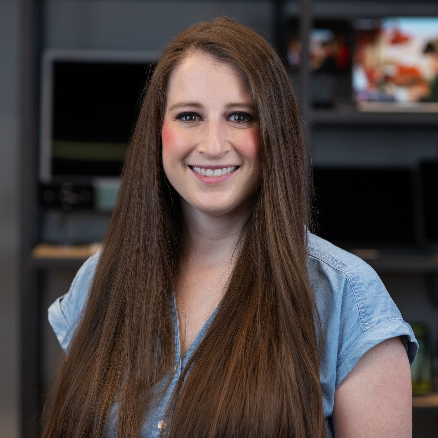 Professional headshot of person in light blue top with long straight hair against dark background.