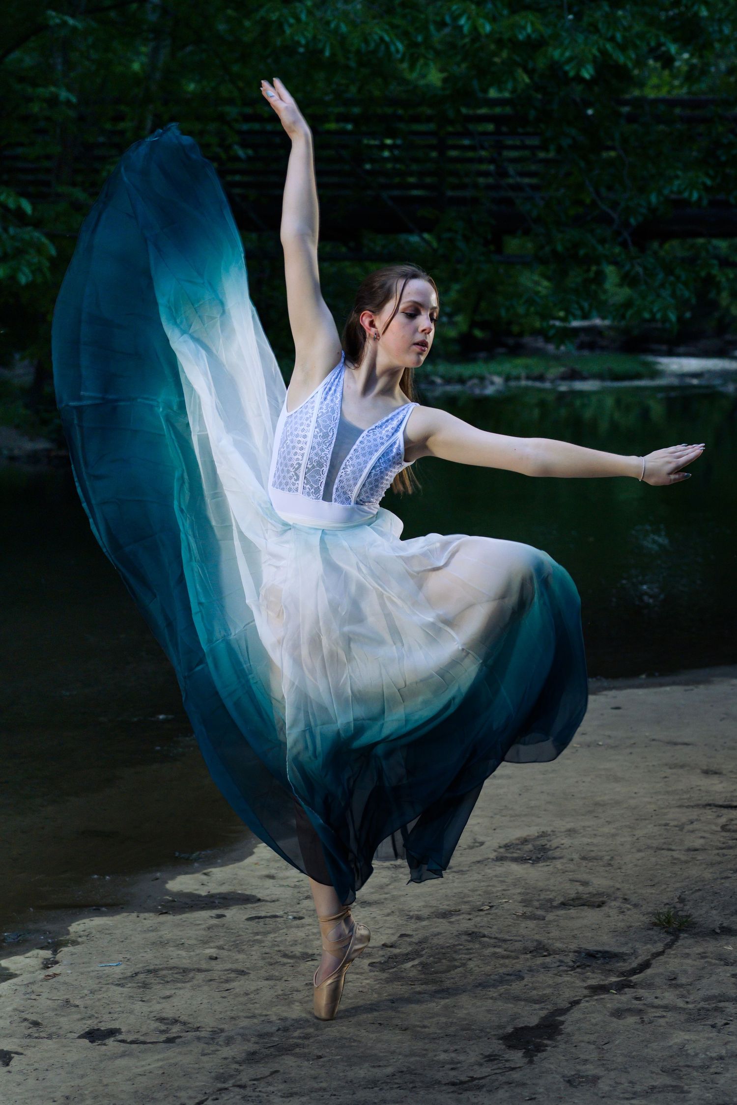 A graceful ballerina leaps in a flowing teal and white gown on pointe shoes near a serene forest pond.