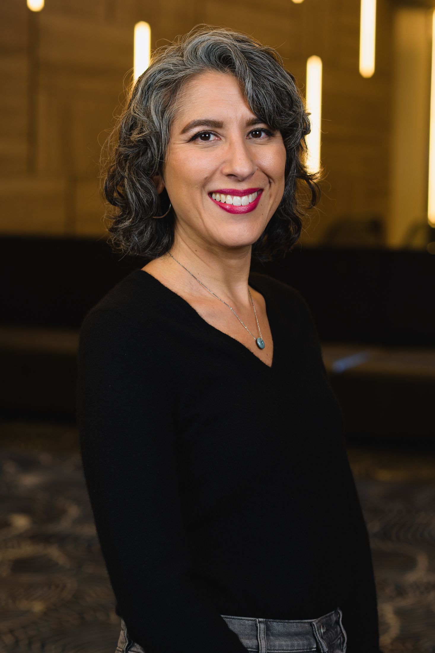 Business headshot showing someone in black attire with styled hair against warm lighting.