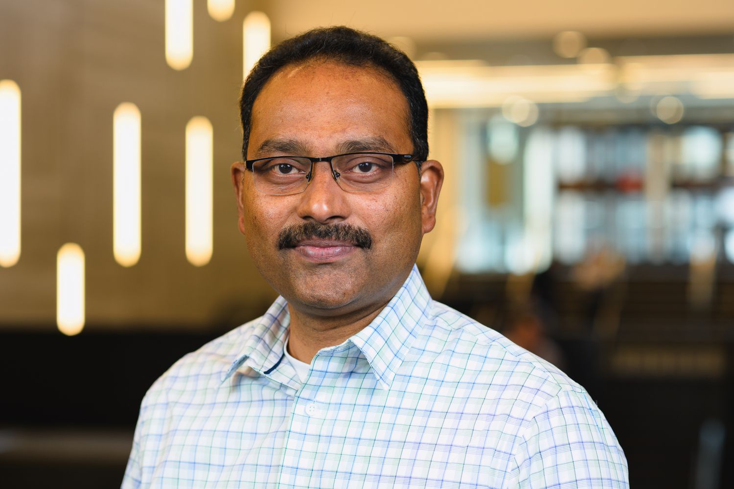Professional headshot of person in checkered shirt against modern office background with vertical lighting.