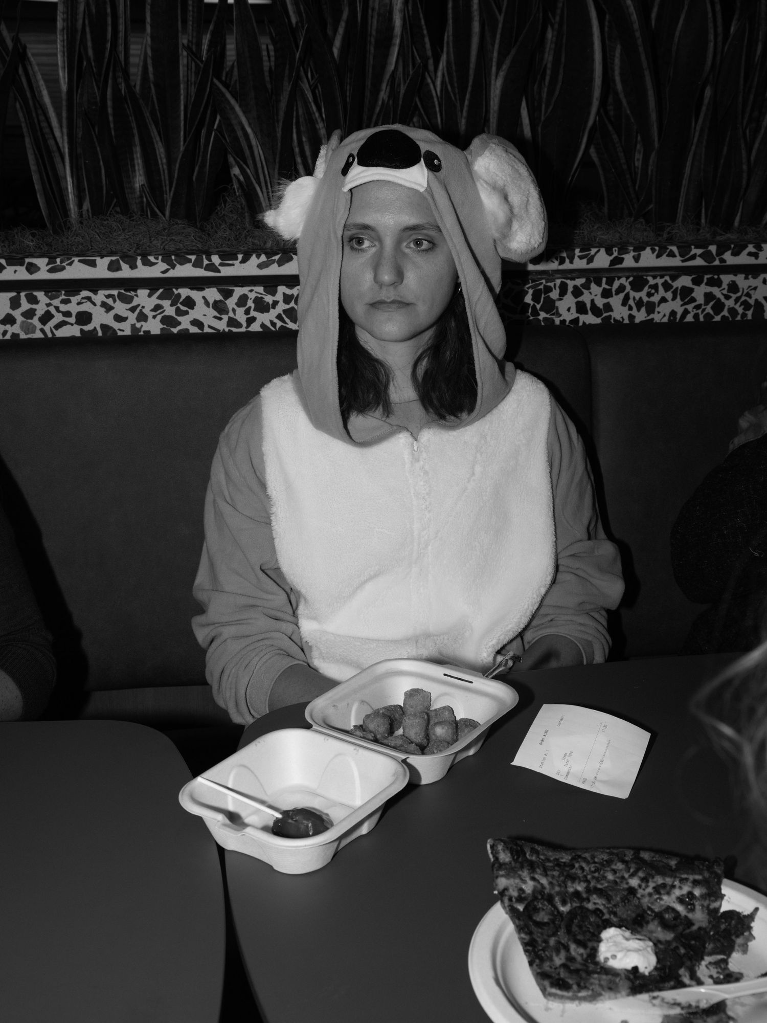 A person wearing a white animal onesie sits at a table with food in black and white photography.