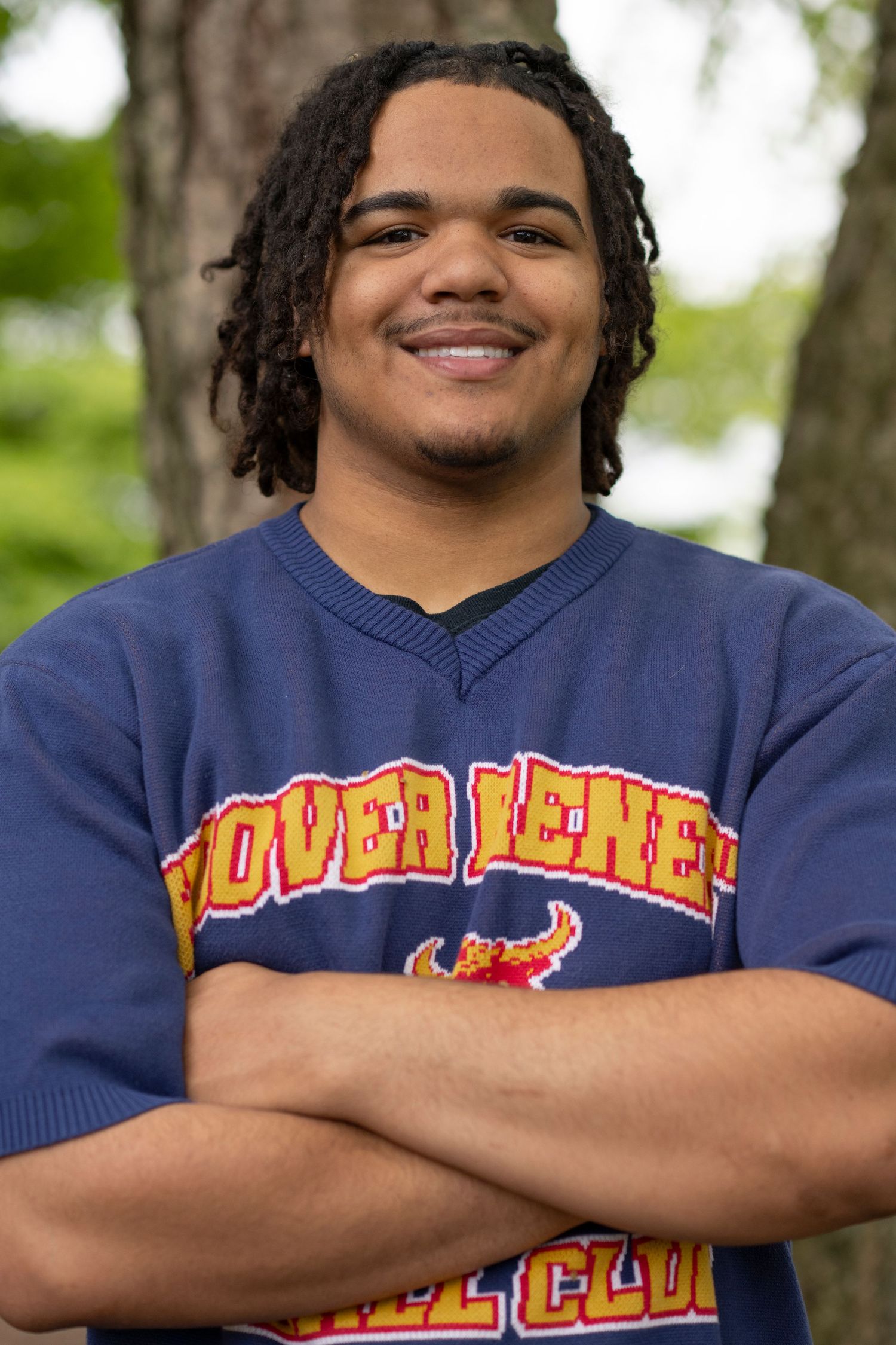 Student in navy blue sweatshirt with dreadlocks smiles warmly with arms crossed against blurred outdoor background.