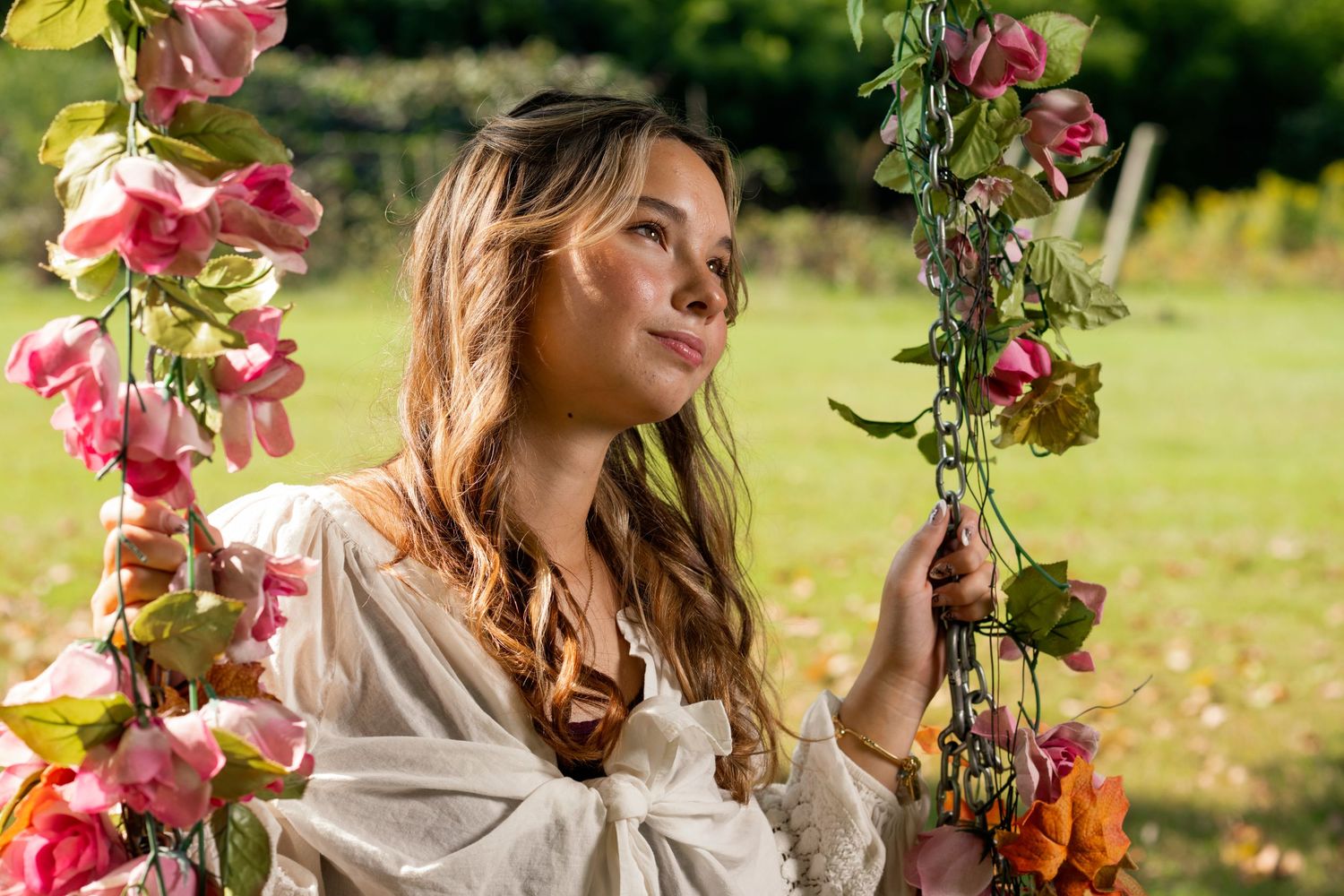 A woman in a white dress poses among pink flowers and greenery in a sunny garden setting.