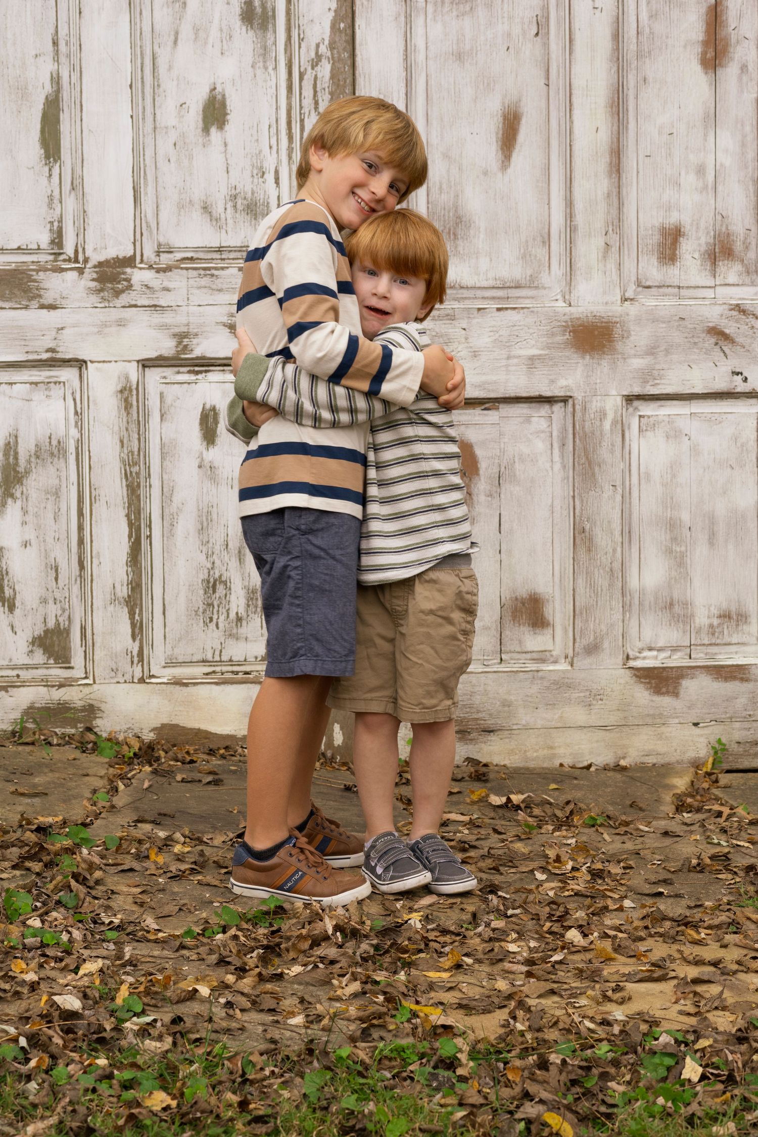 Two young children in striped clothing hugging against weathered white door background.