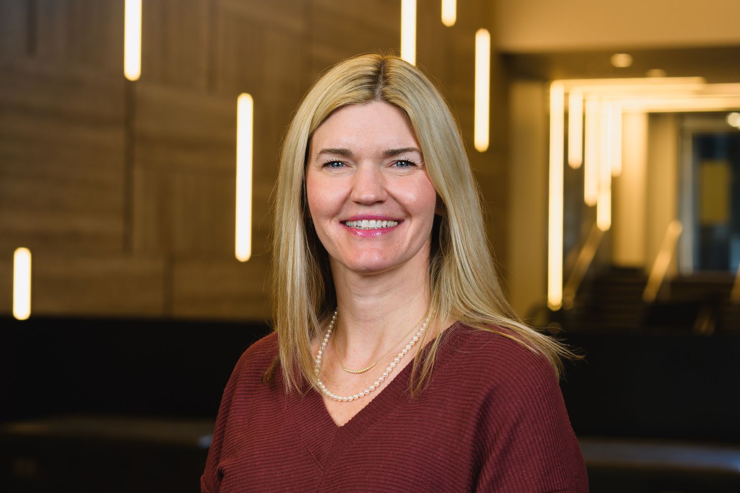 Professional headshot of someone in burgundy top with blonde hair against warm office lighting.