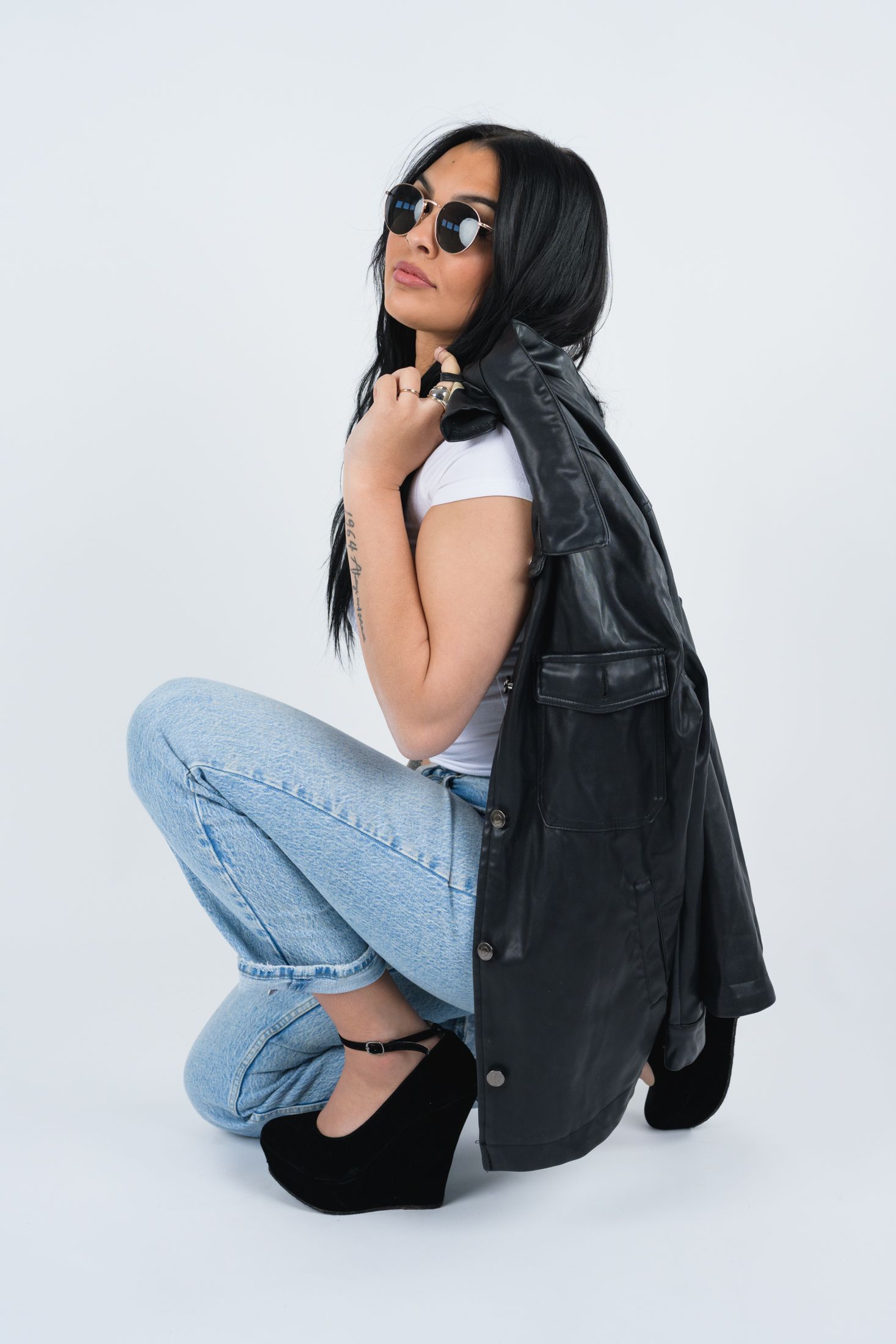 Model in denim and leather poses with sunglasses in minimalist studio setting.
