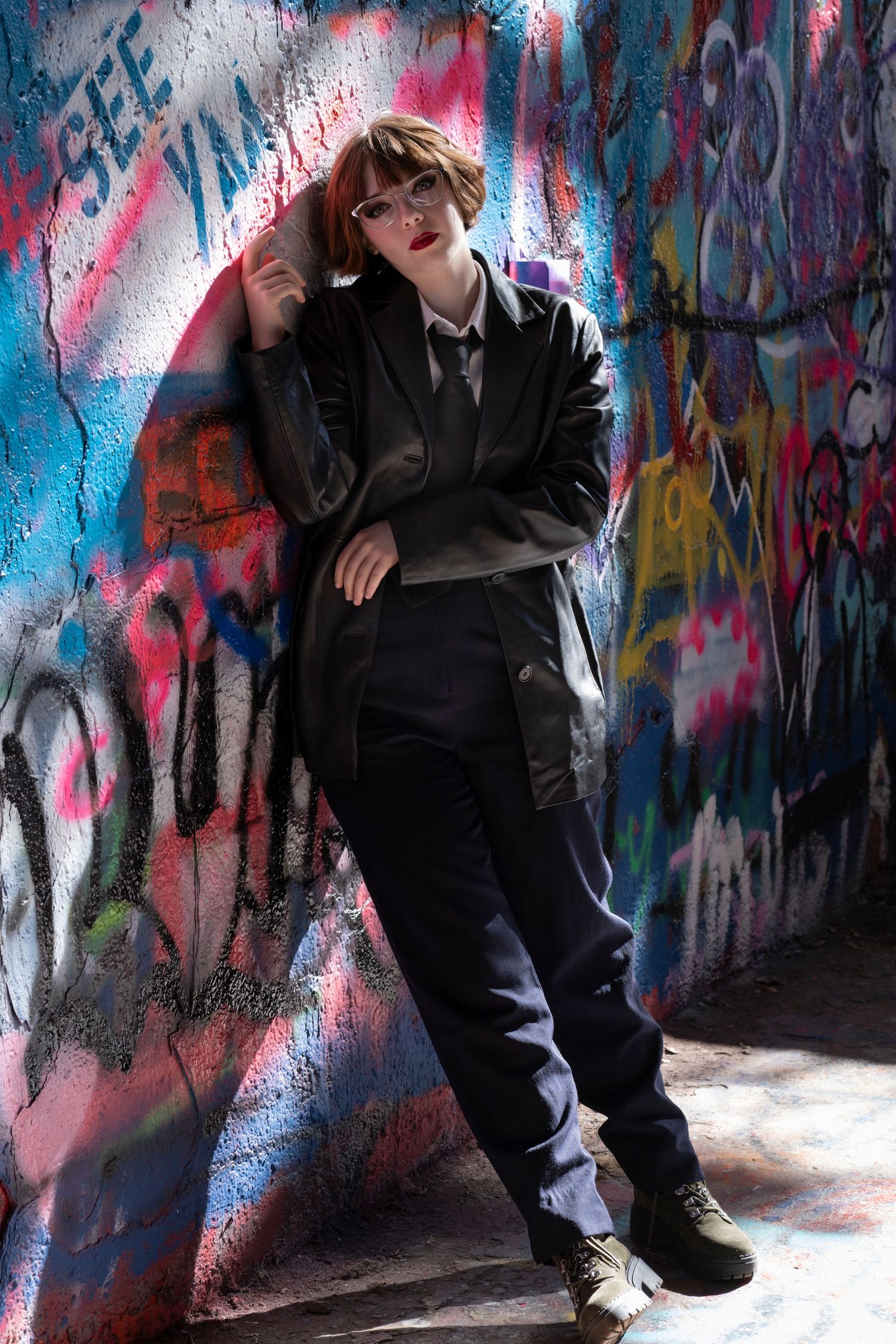 A person in dark clothing poses against a vibrant graffiti wall with pink and blue spray paint designs.