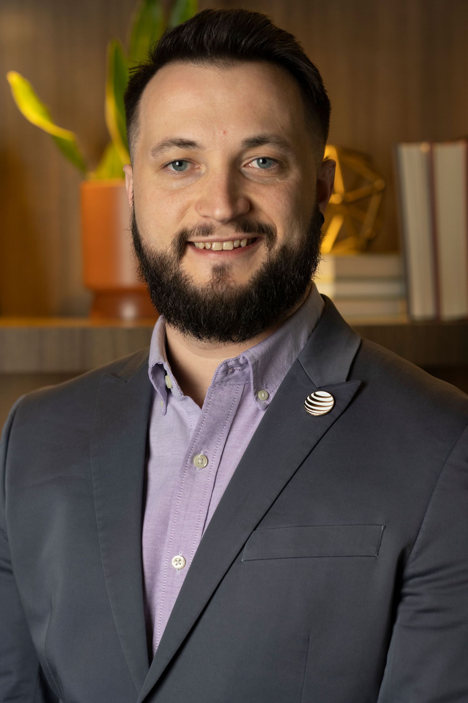 Professional headshot of person in purple dress shirt and dark blazer with AT&T logo against warm office background.