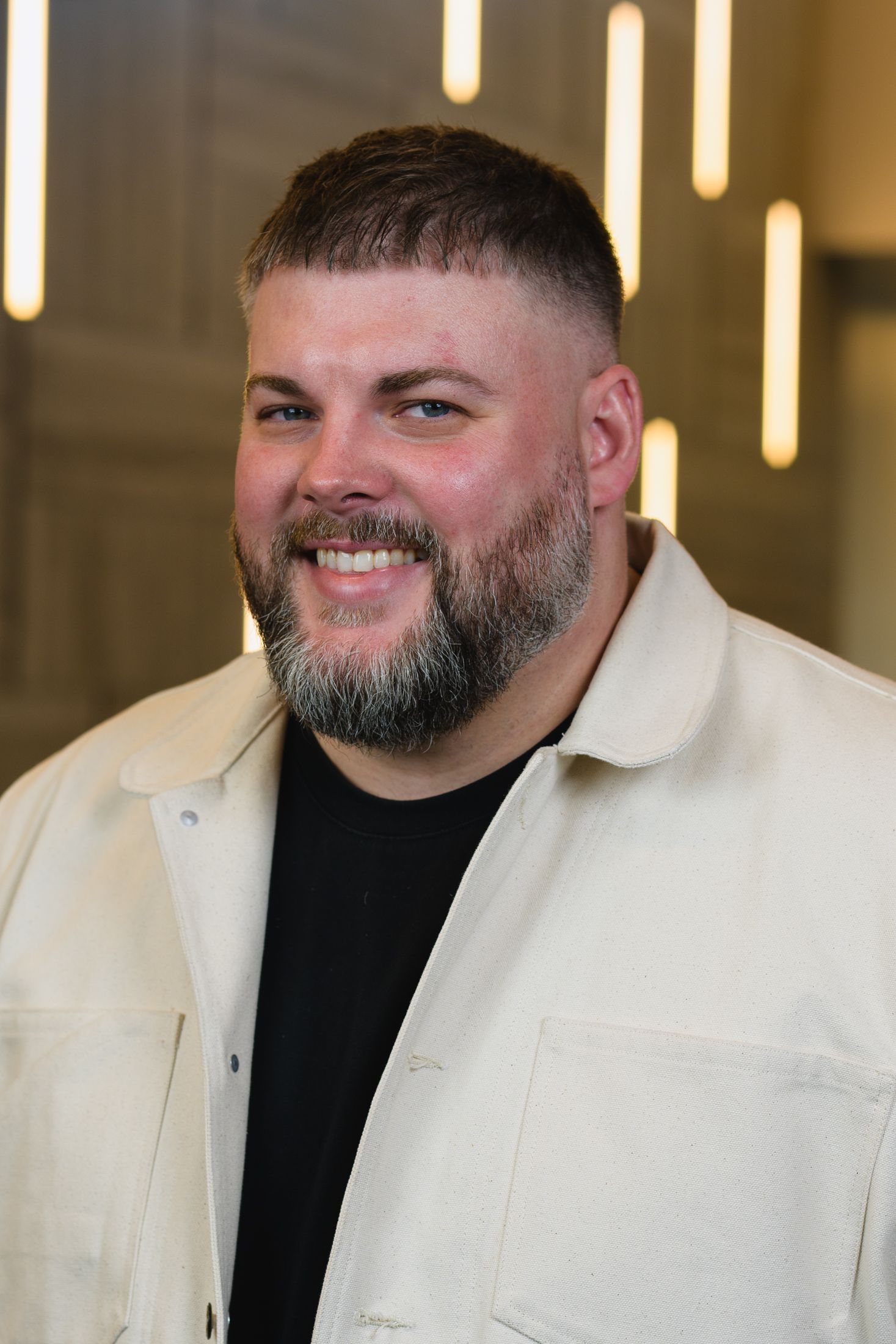 Professional portrait of person in cream jacket and black shirt against modern office lighting.