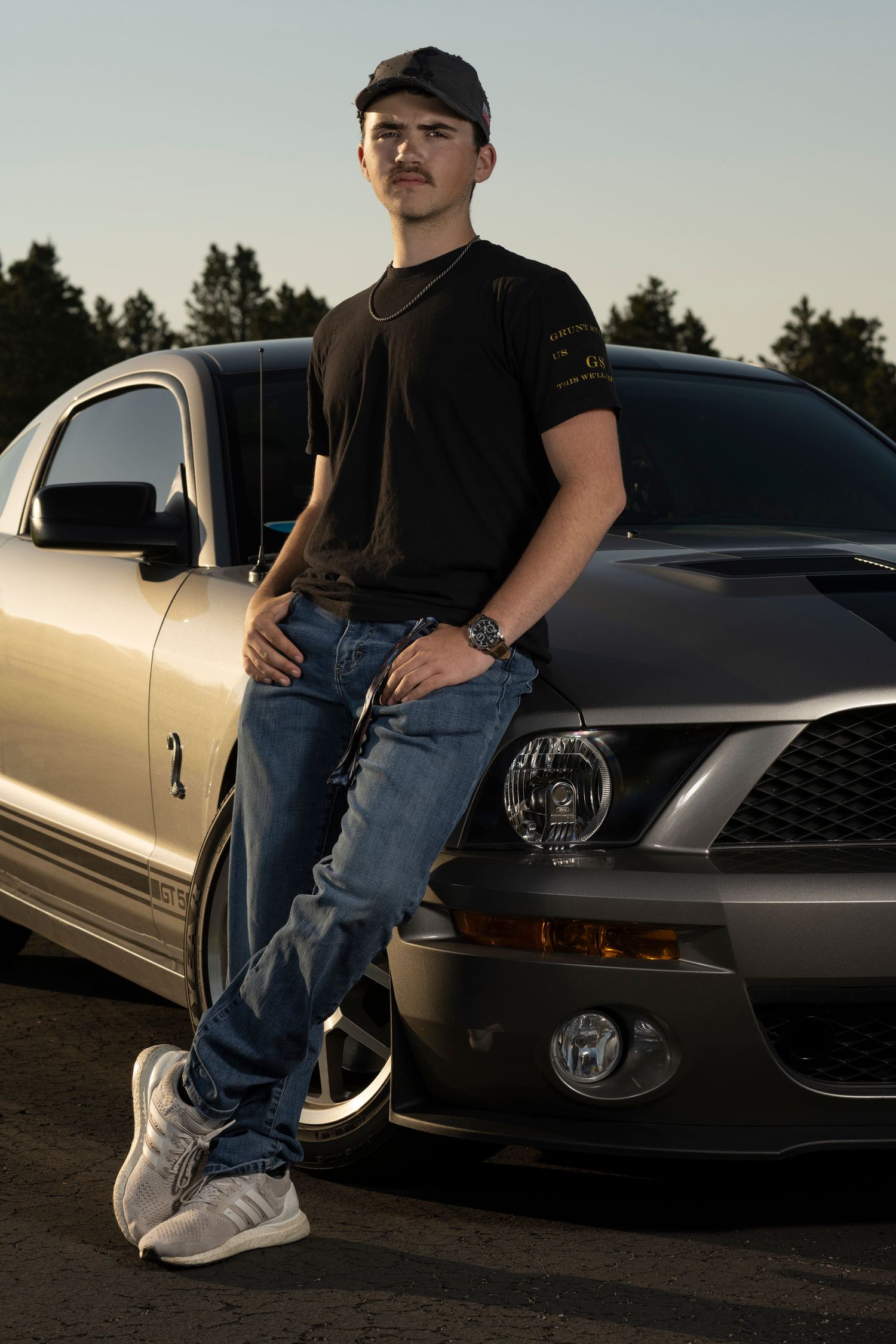 A person in jeans and sneakers poses with a silver sports car at sunset, showing various angles of the vehicle.