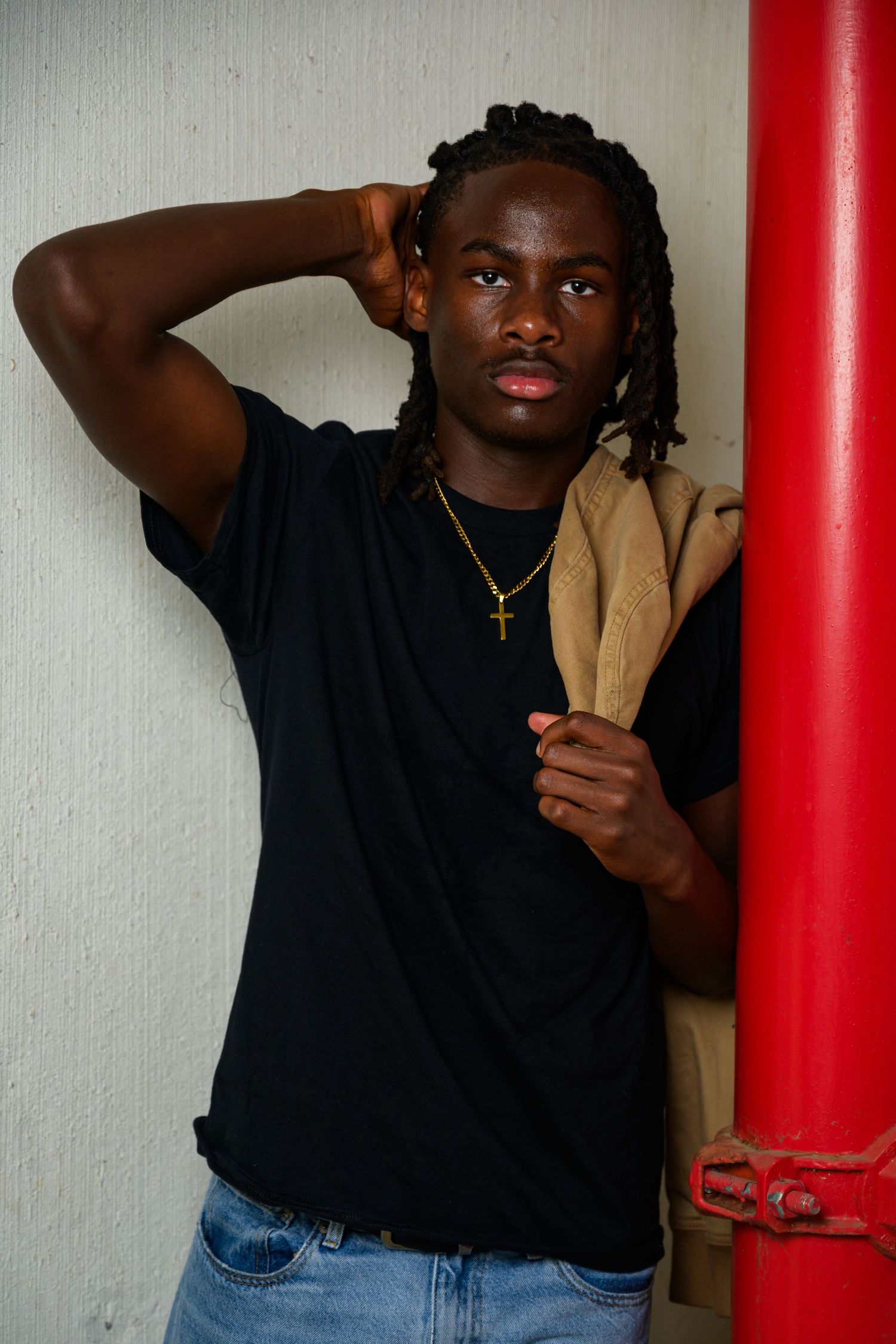 Young Black man with braids wearing black t-shirt and gold cross necklace, leaning against wall holding tan jacket.