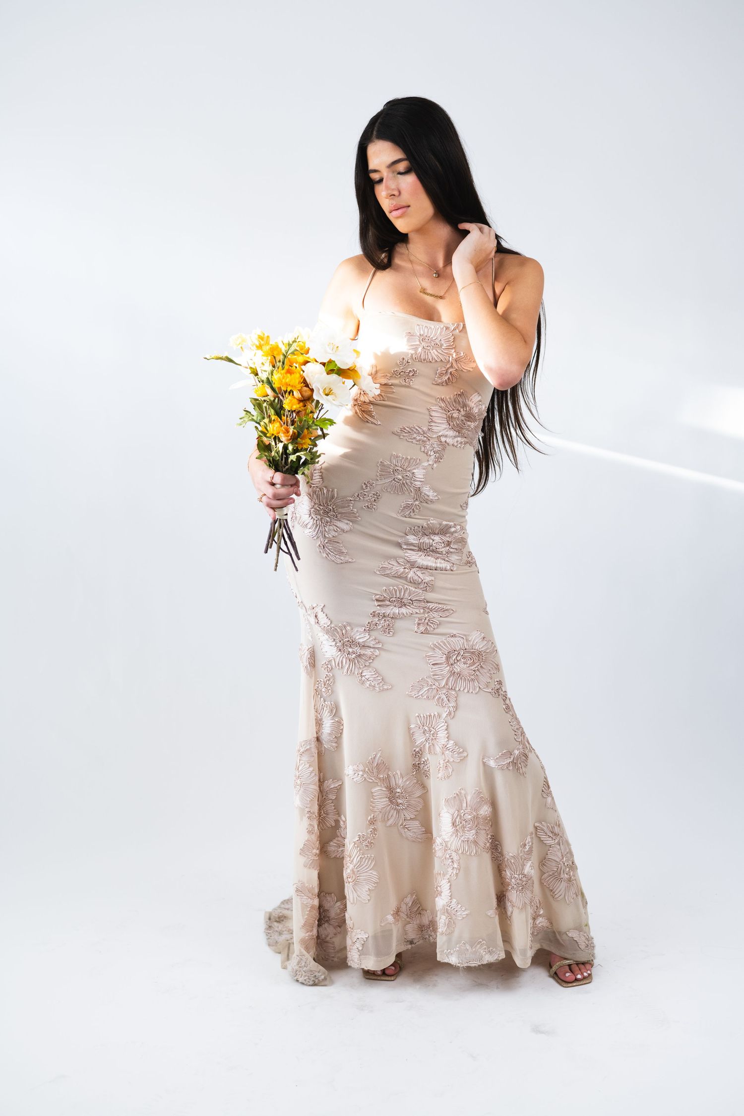 Woman in beige floral dress poses with yellow flowers against white backdrop.