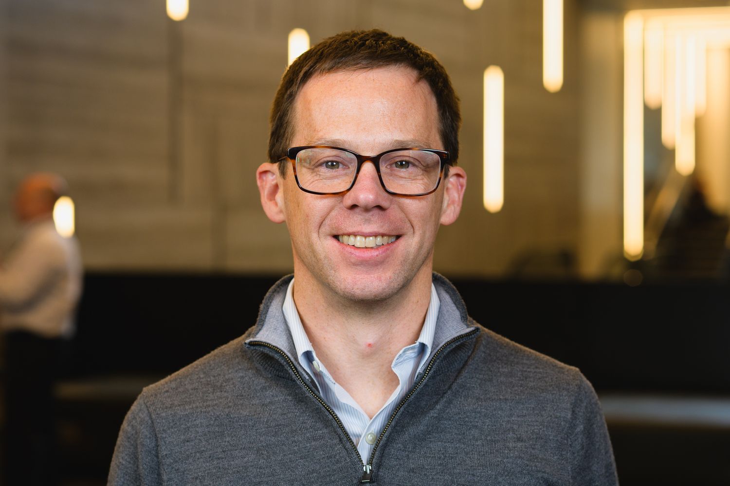 Professional headshot of person in glasses and blue dress shirt against office background with modern lighting.