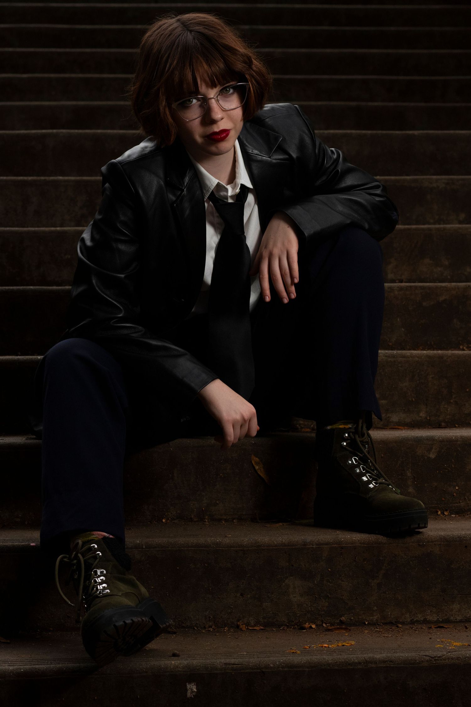 A person in a dark suit and glasses sits contemplatively on wooden steps in moody lighting.