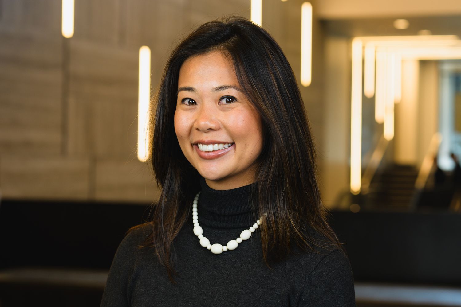 Professional headshot of person wearing white necklace against modern office background.