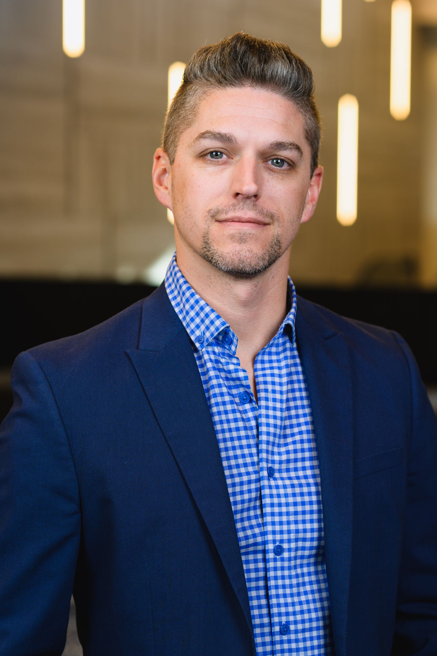 Business portrait showing a person in a navy suit with blue gingham shirt against warm lighting.