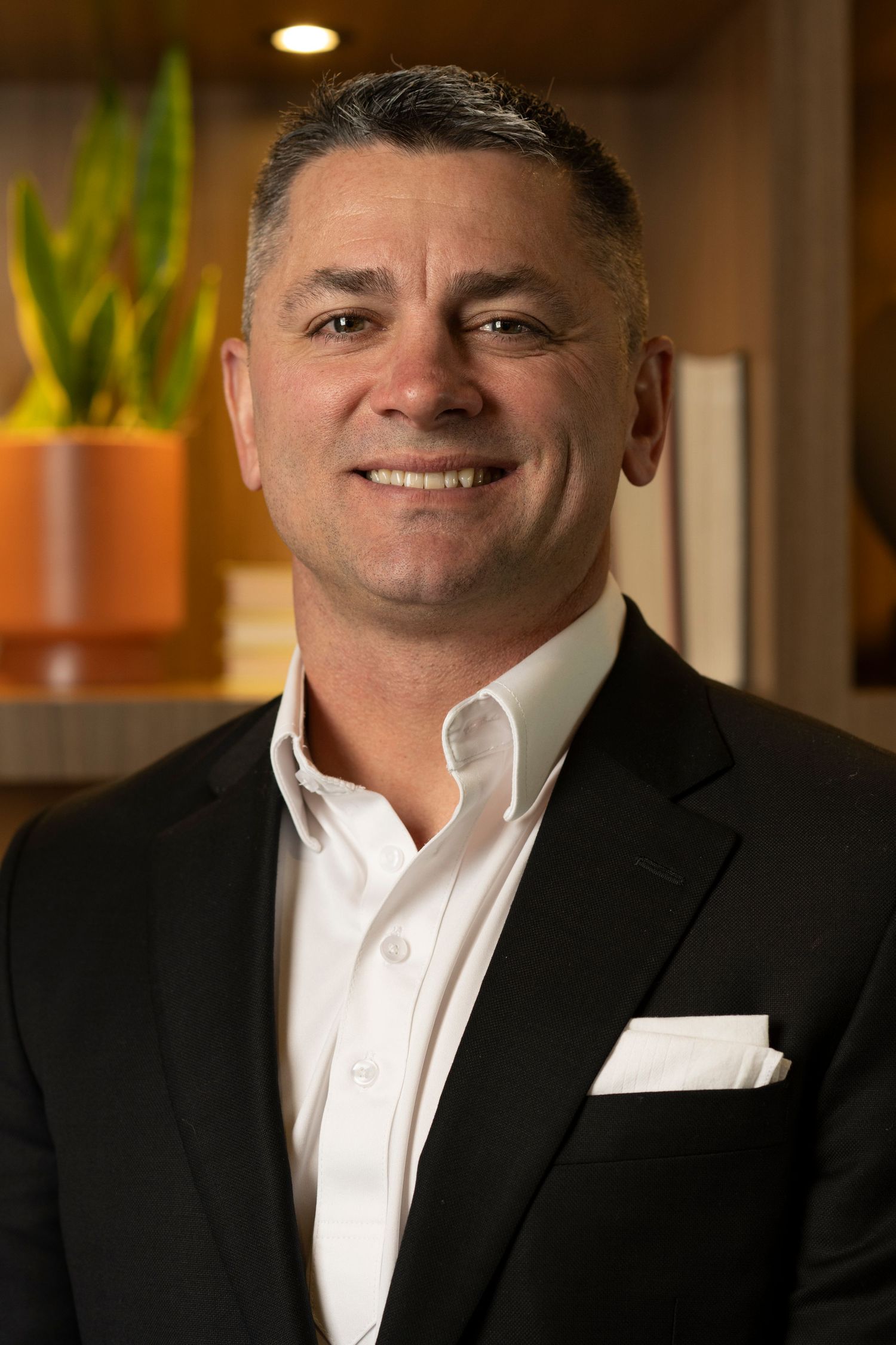 Professional portrait of person in white dress shirt and black suit jacket against warm-lit office background.