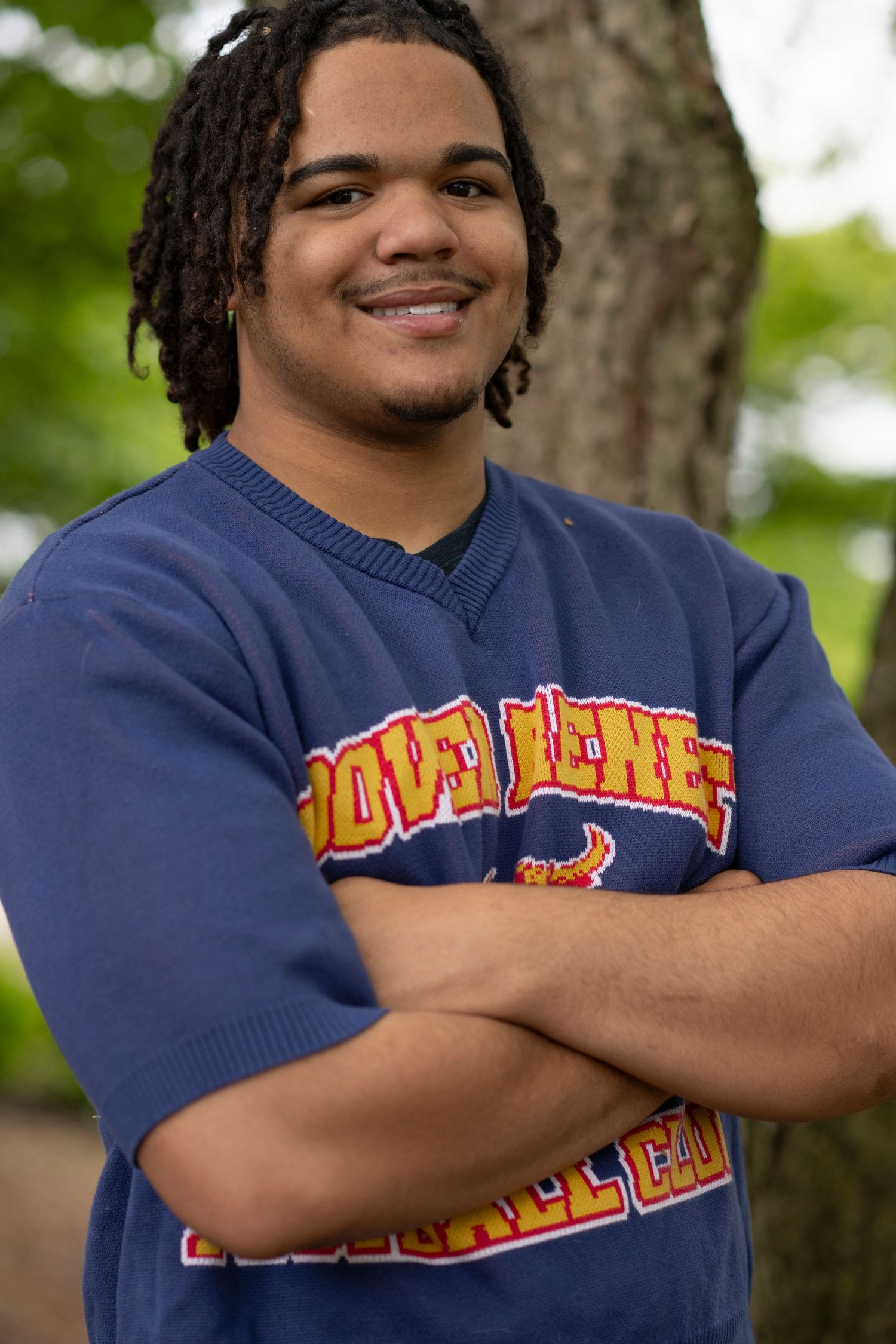 A person wearing a navy blue sweatshirt with orange text smiles while posing outdoors against a blurred green background.