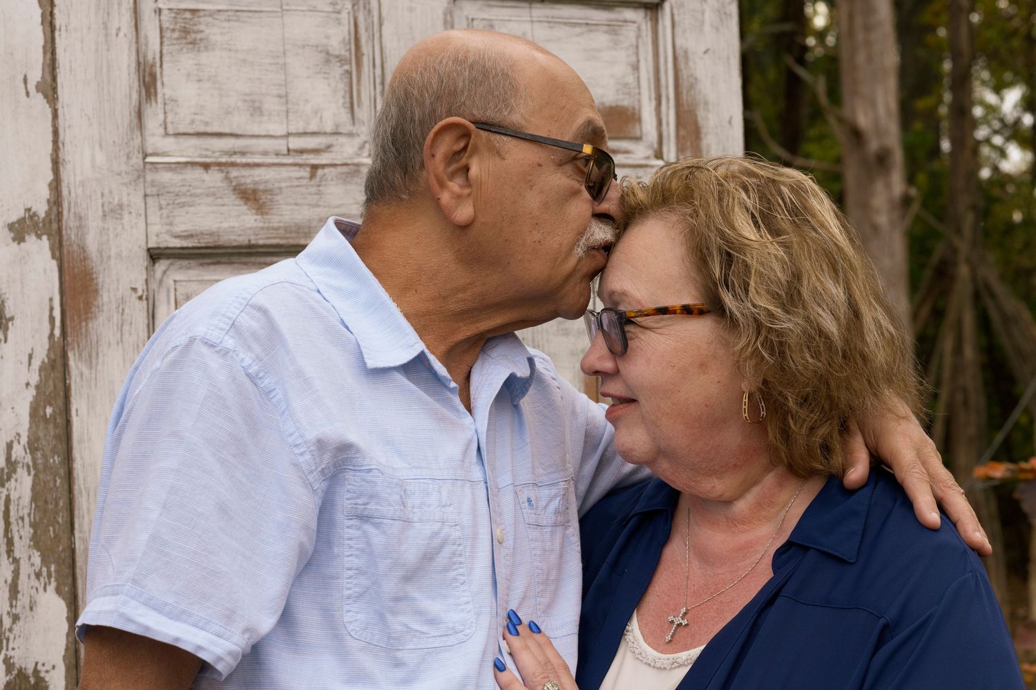An elderly couple embraces lovingly while sitting outdoors against a white wooden backdrop.