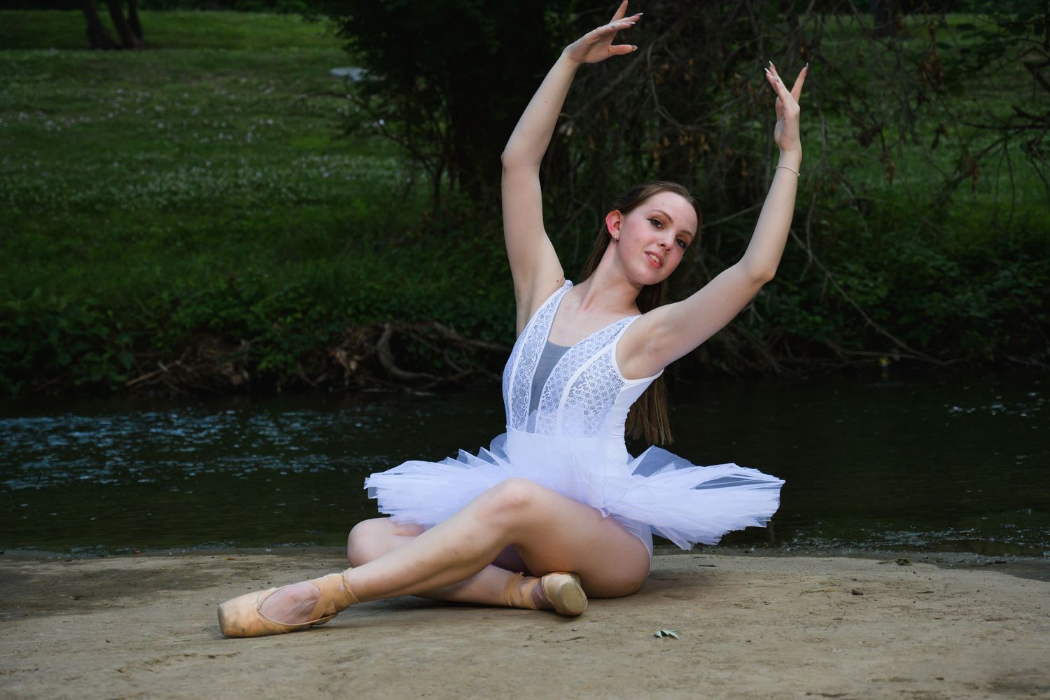 A young ballerina in a white tutu performs an elegant pose outdoors near a creek, wearing pointe shoes.